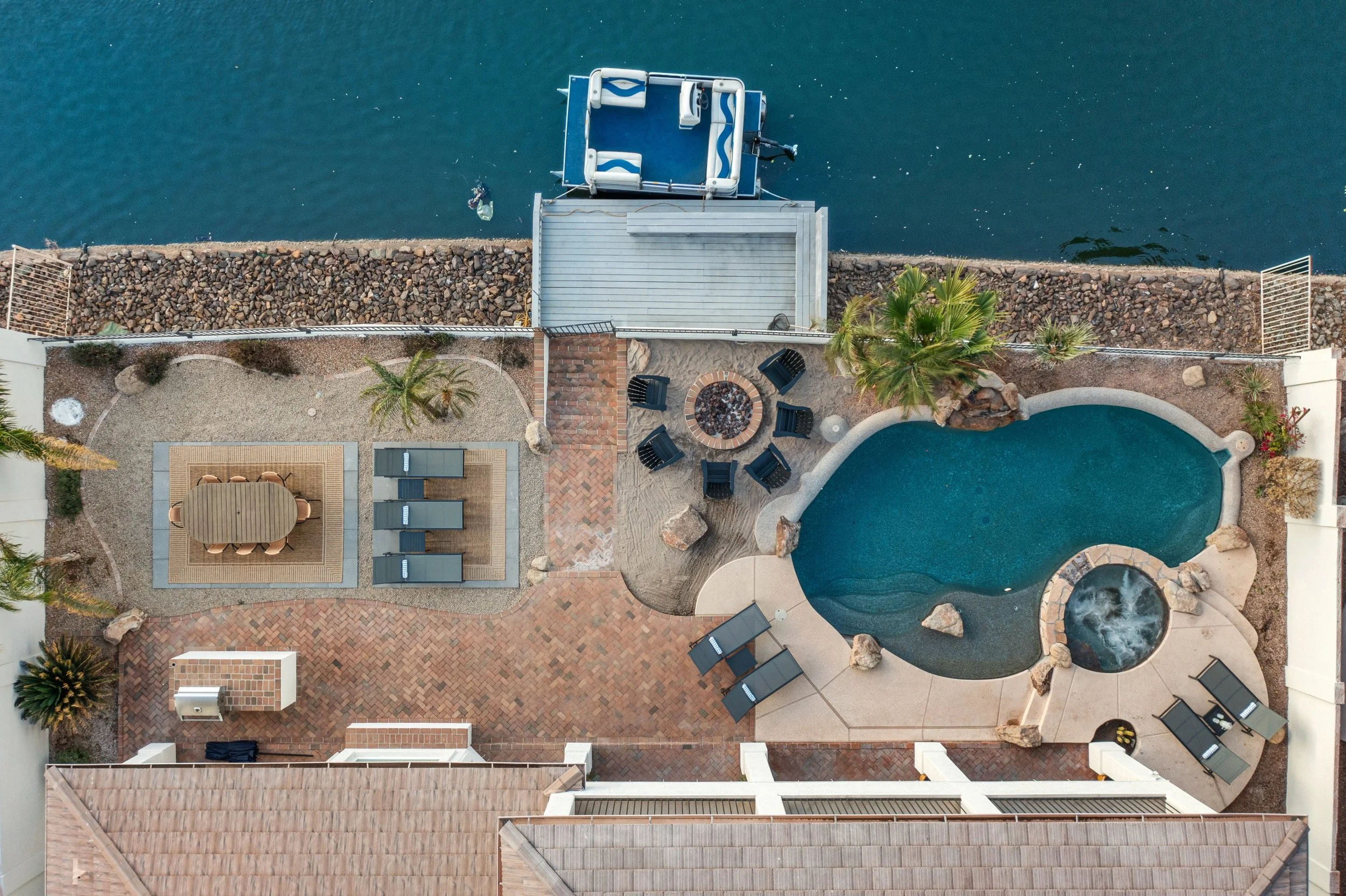 Aerial view of a backyard with a pool, fire pit, patio furniture, palm trees, and a dock with a boat on the water.
