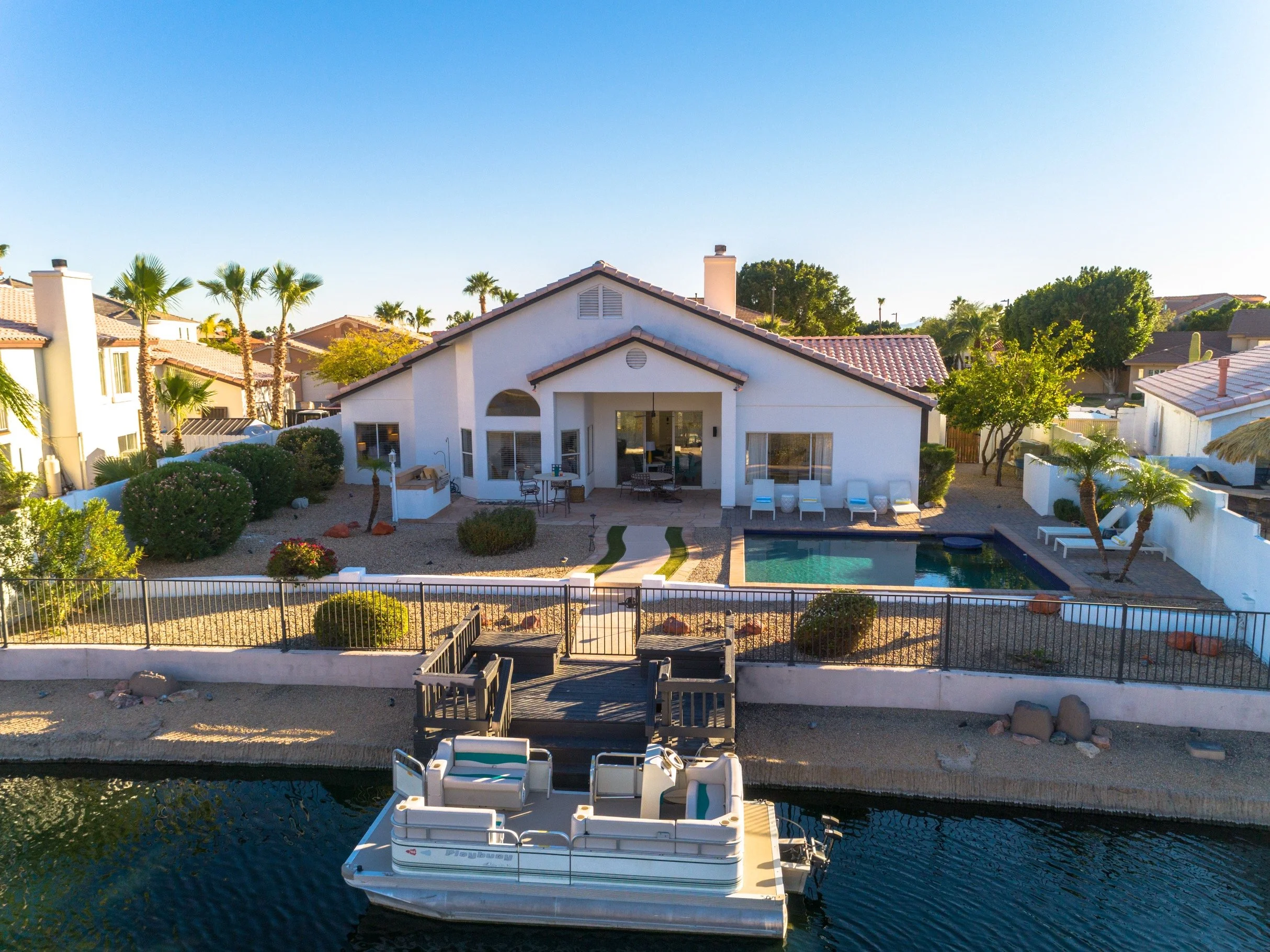 Backyard with a swimming pool, outdoor furniture, and a house with a patio, surrounded by palm trees and other greenery in a sunny neighborhood.
