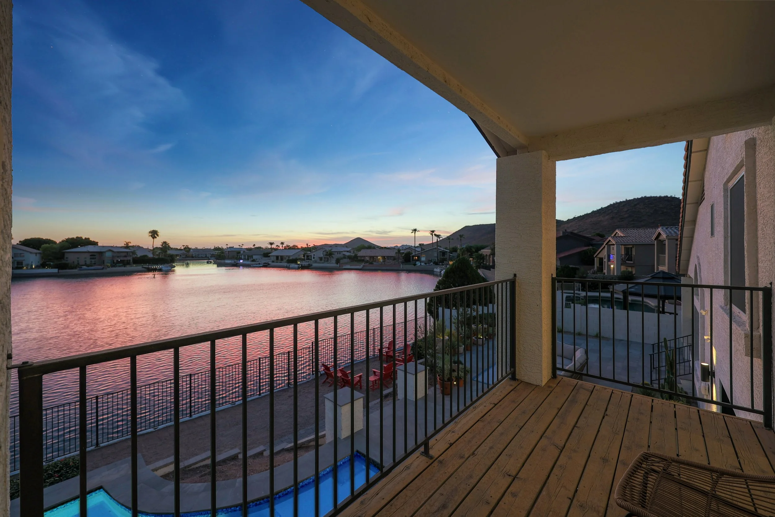 Balcony view of a neighborhood with houses along a waterway, hills in the background, and a sunset sky with clouds.