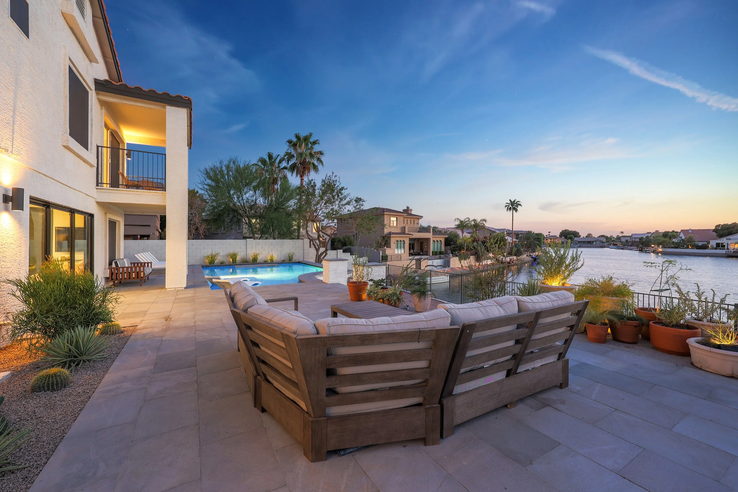 A backyard patio with outdoor seating, including a curved sofa, a lounge chair, and a picnic table with chairs. There is a small pool, potted plants, and desert landscaping. The house is two stories with exterior lights, and the scene overlooks a can