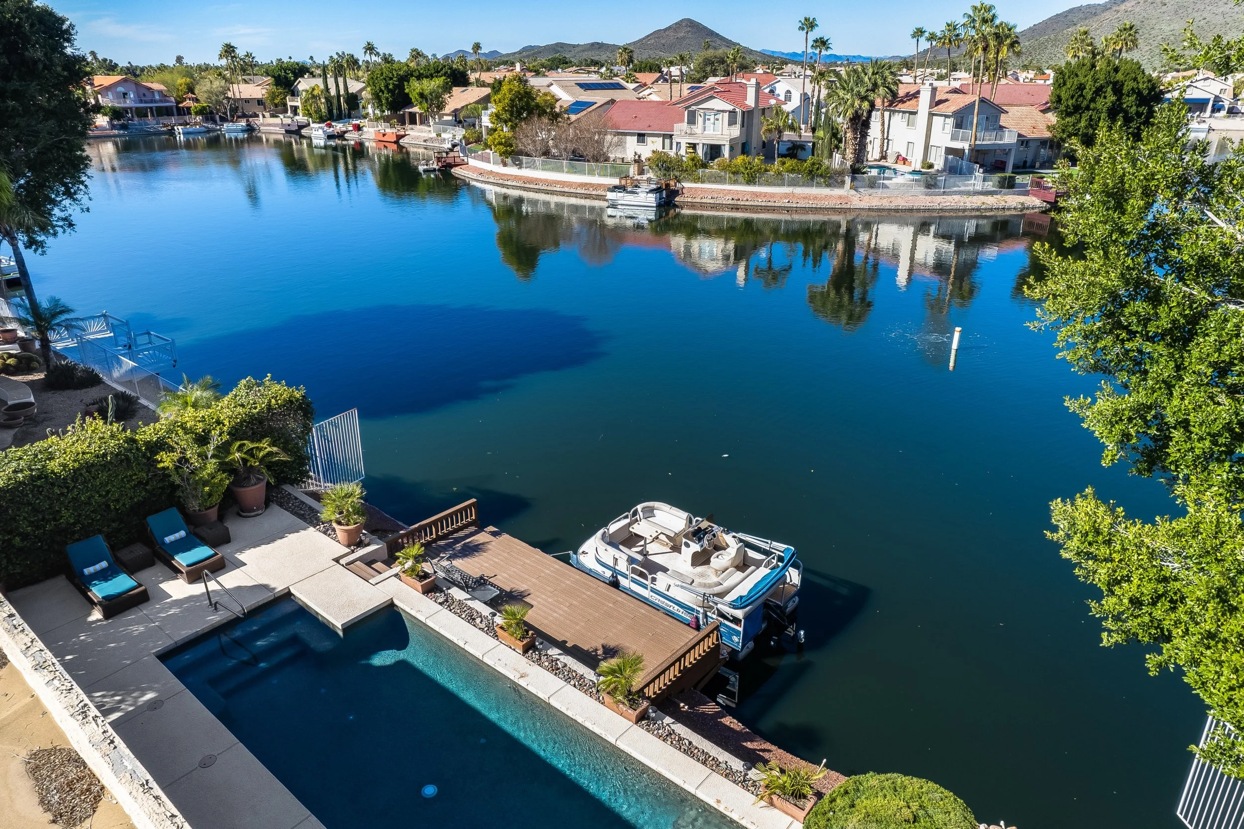 A backyard with a swimming pool, boat dock, and waterfront houses, with palm trees and mountains in the background.