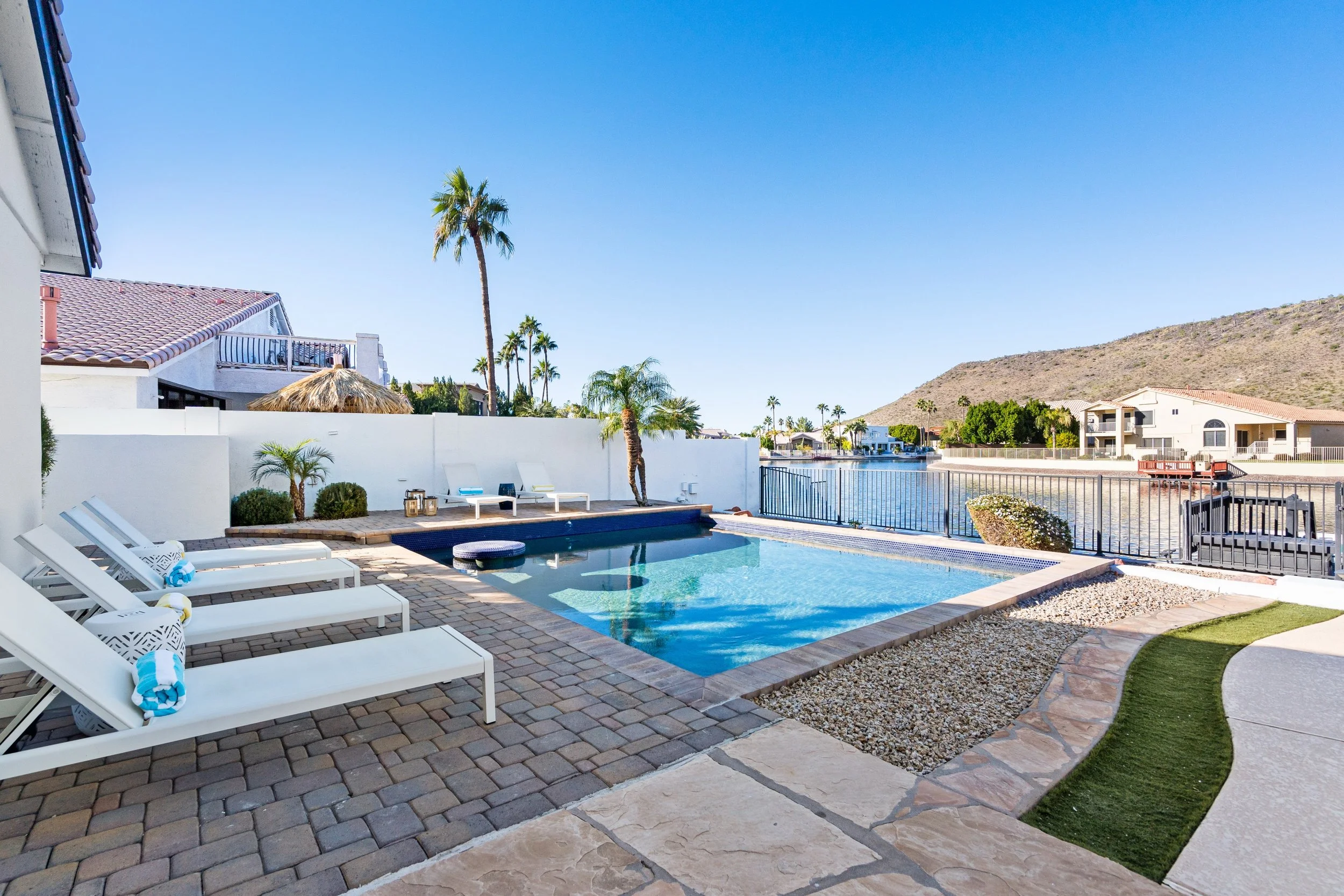 A backyard with a swimming pool, lounge chairs, palm trees, and a view of a canal with houses and mountains in the background.