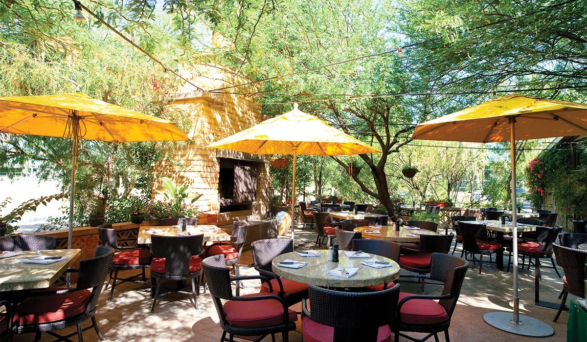 Outdoor restaurant dining area with several tables, black chairs with red cushions, yellow umbrellas, surrounded by trees and greenery