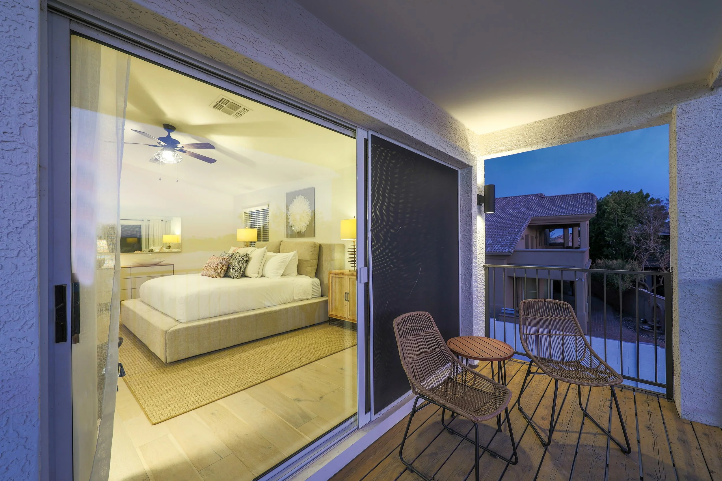 Balcony view at dusk with outdoor seating and a sliding glass door leading into a bedroom with a bed and lamps.