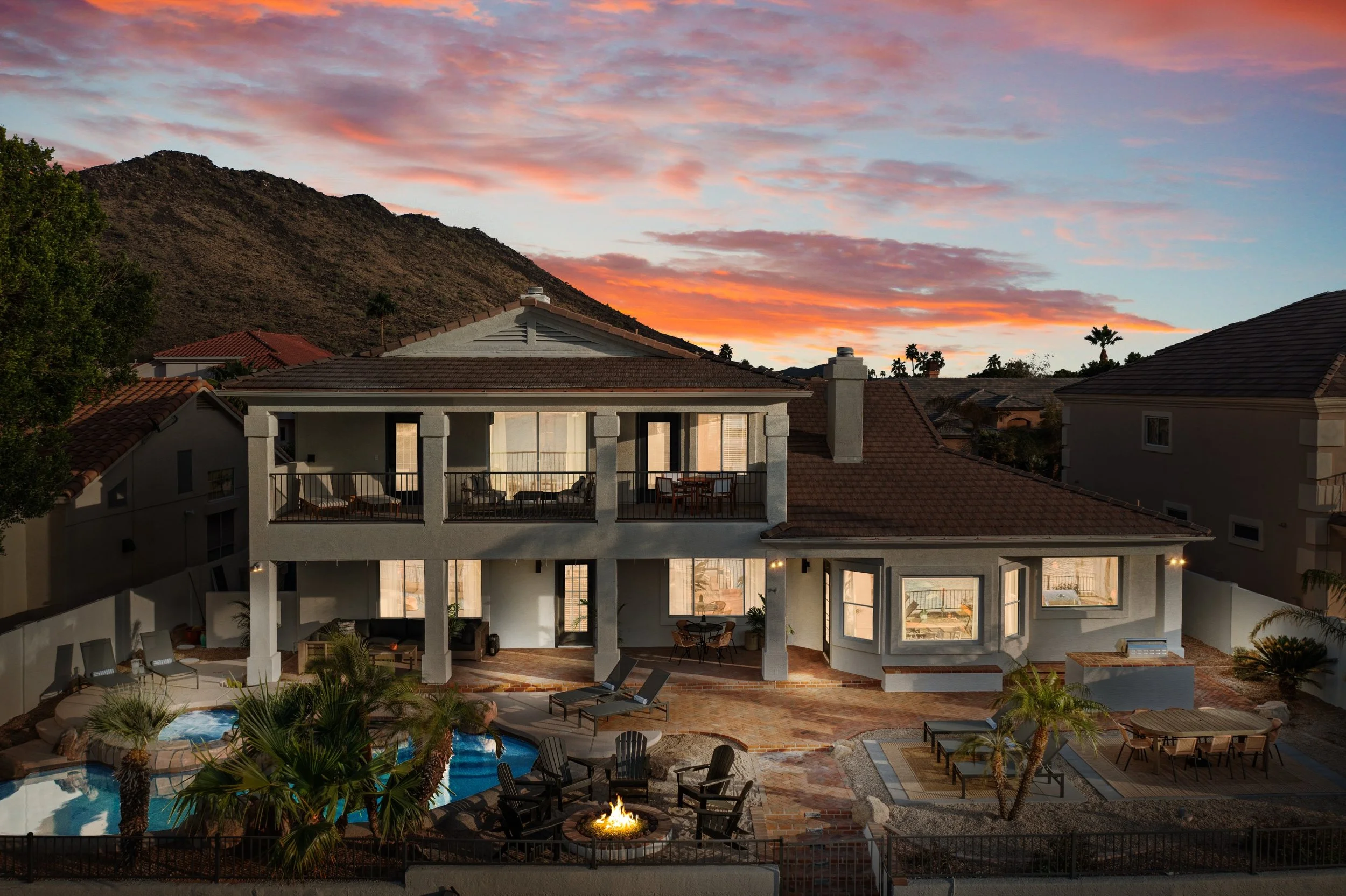 A large modern house with an outdoor swimming pool, patio chairs, a fire pit, and lush landscaping, set against a backdrop of mountains and a colorful sunset sky.