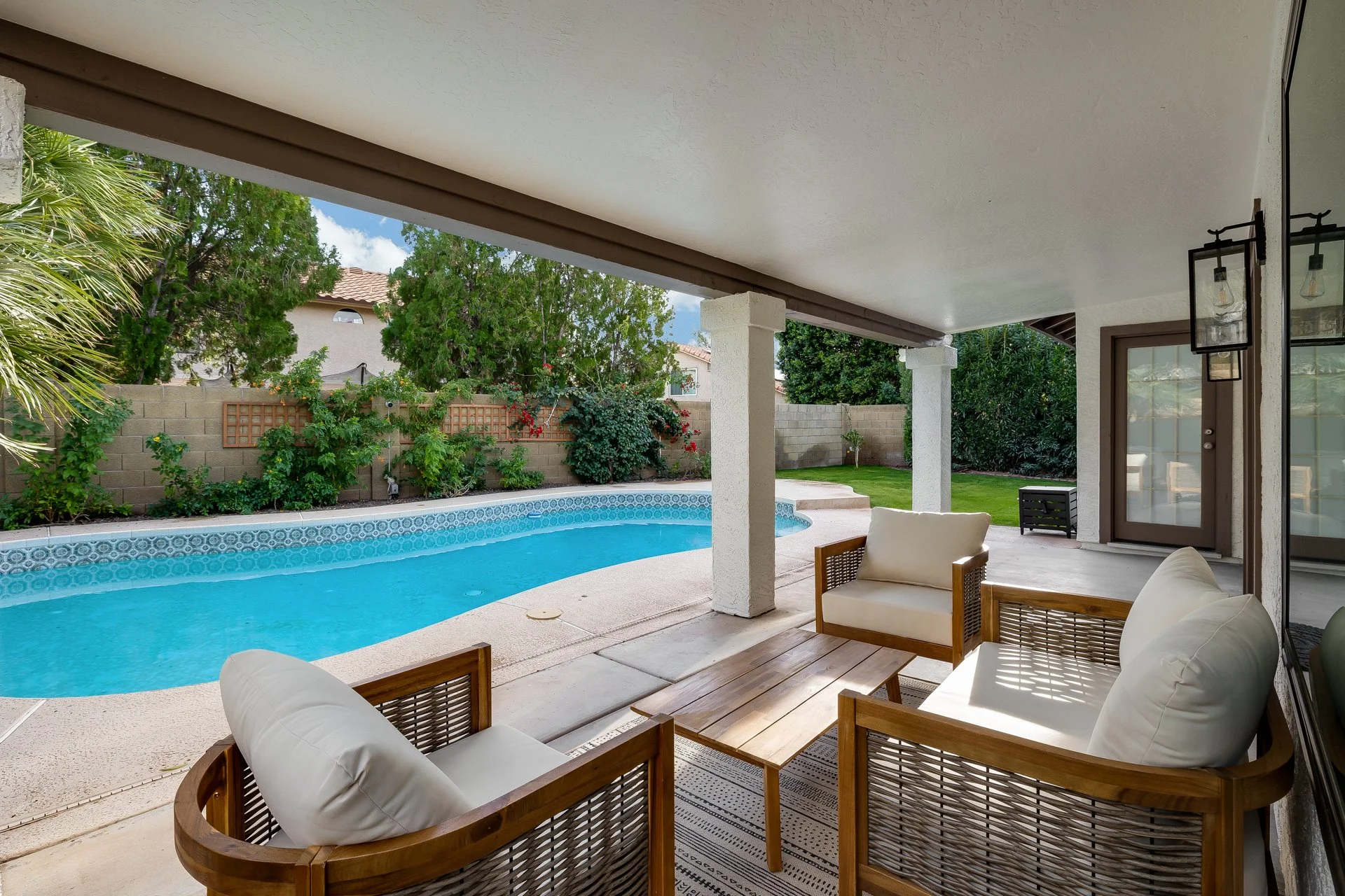 Covered patio area overlooking a backyard pool, with wooden furniture and lush greenery.
