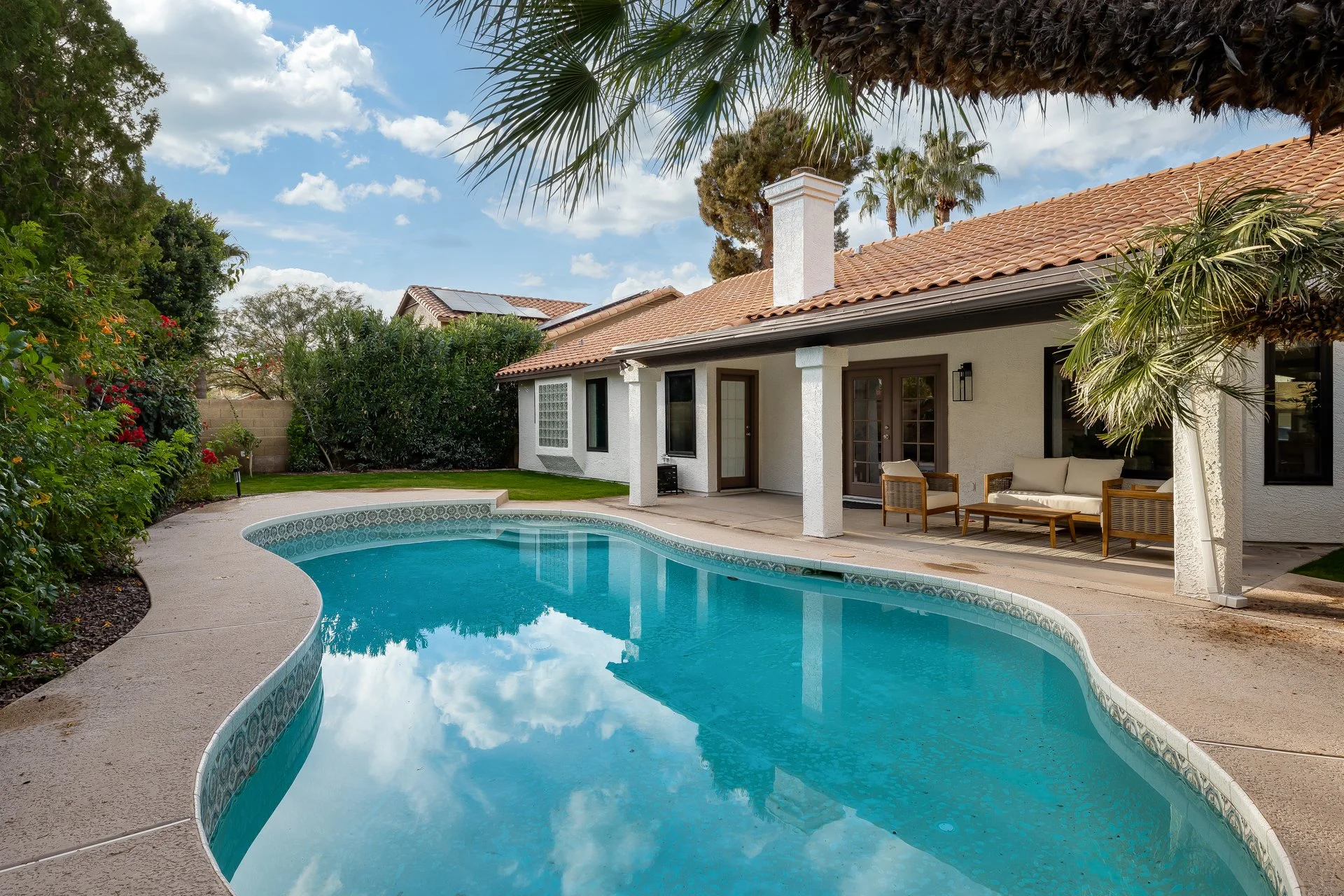 A backyard with a curved swimming pool, surrounded by a concrete deck. There are outdoor chairs and a table on a covered patio area attached to a white house with a tiled roof. The yard has lush green plants and trees, with a brick wall and a blue sky with scattered clouds in the background.