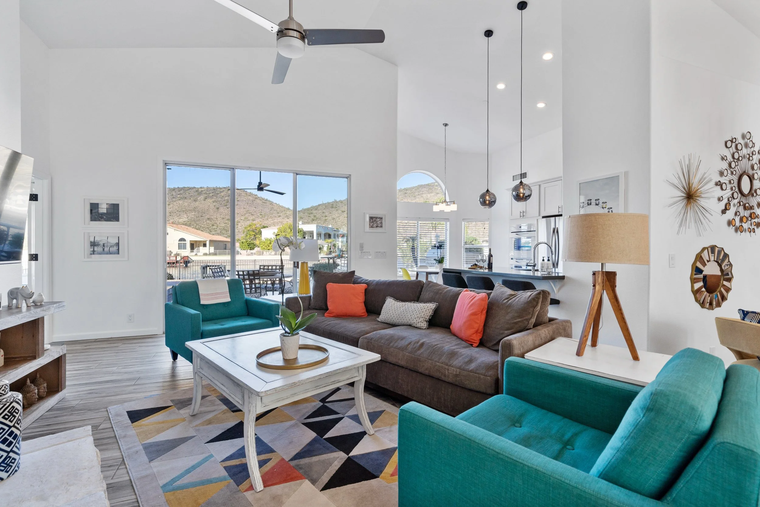 A living room with white walls and a large window showing a mountainous landscape outside. The room is decorated with colorful furniture including teal armchairs, a brown sofa with orange and gray pillows, and various modern art and decorative items.