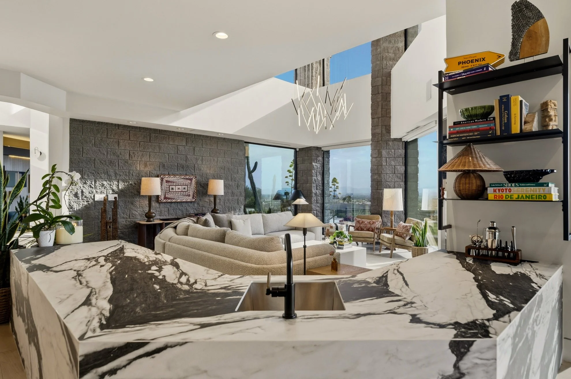 Modern open-concept living space with a marble kitchen island, beige sectional sofa, large windows, brick accent wall, and contemporary lighting fixture.