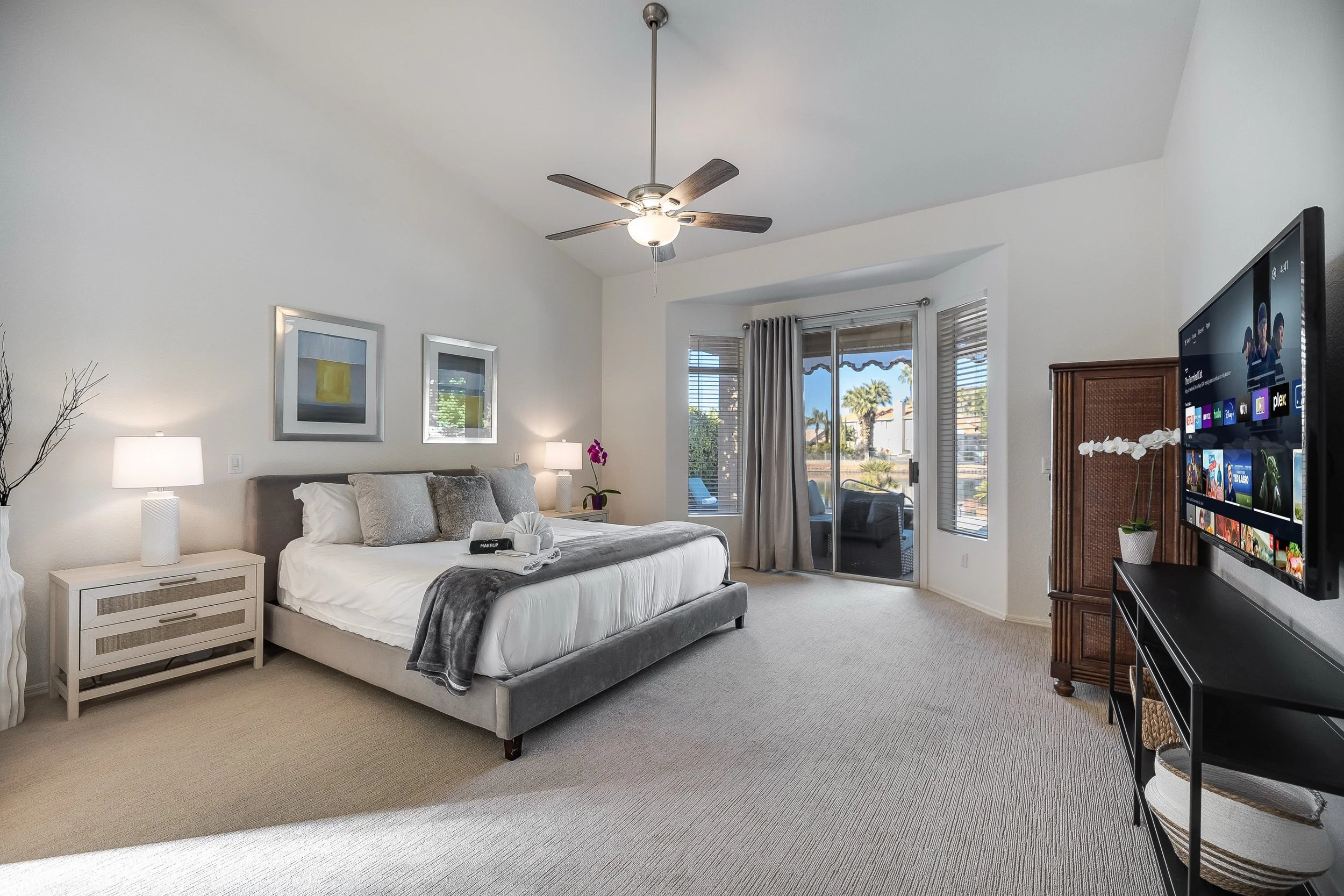 A bedroom with a bed, nightstand, lamps, wall art, a ceiling fan, and a sliding glass door leading to a patio with outdoor furniture and palm trees.