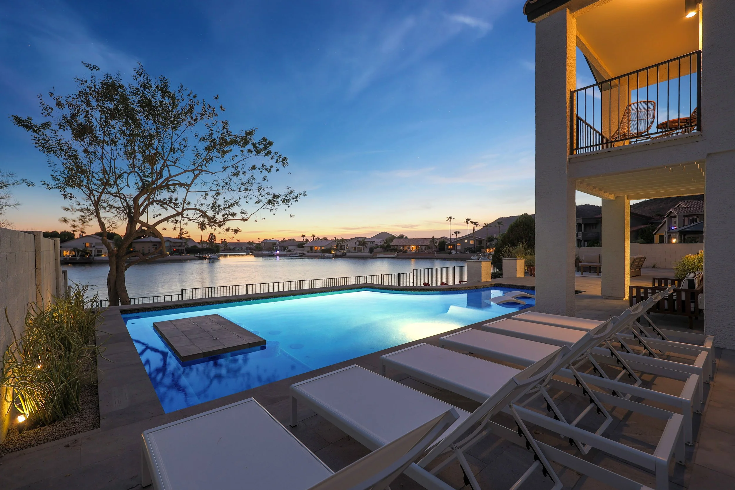 A backyard pool area at dusk with lounge chairs, a tree, and a view of a lake and houses in the distance, under a clear sky.