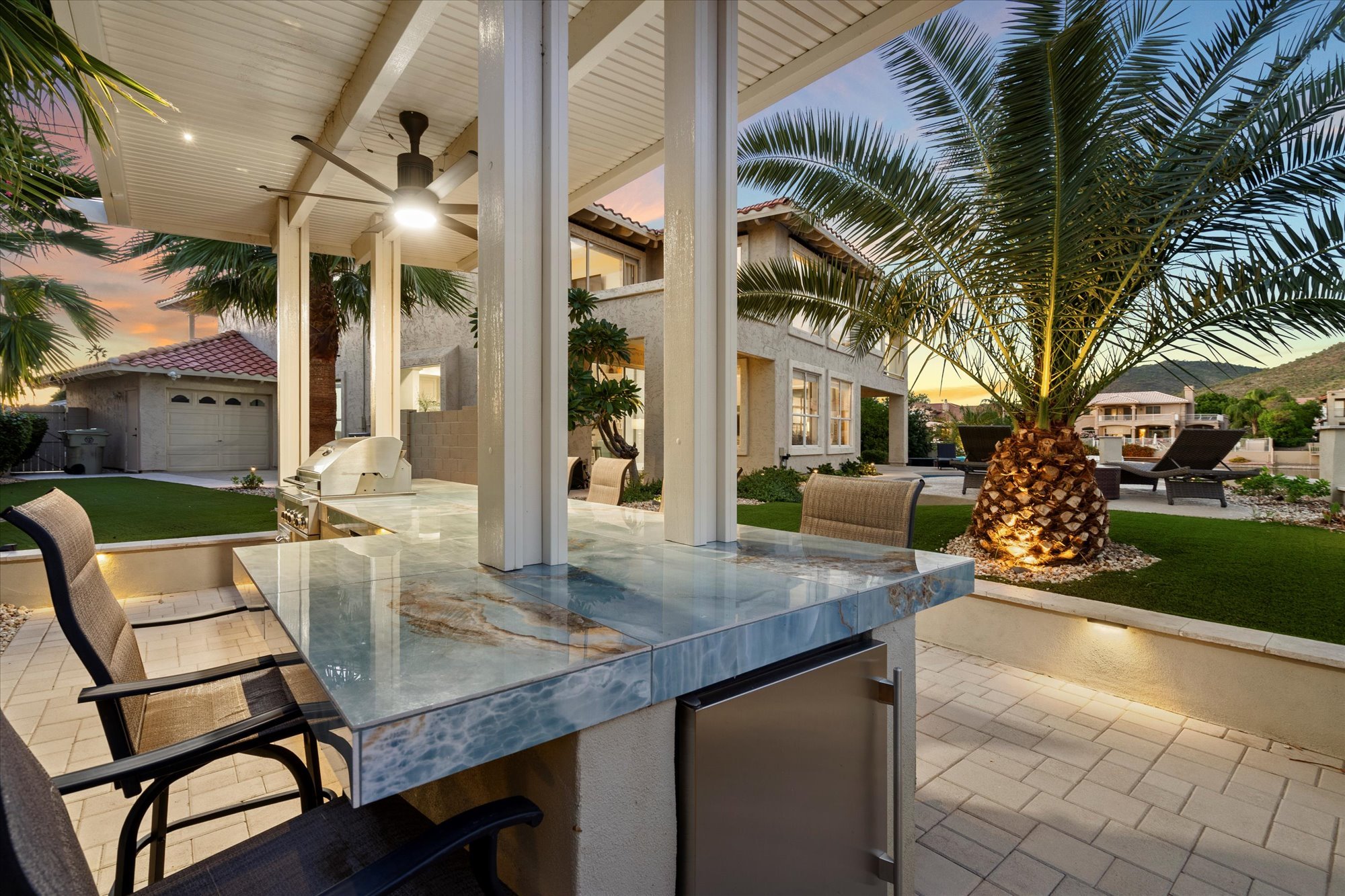 Outdoor patio area with a marble table, surrounded by chairs, an outdoor grill, palm trees, and chairs, with houses and a hillside in the background during dusk.