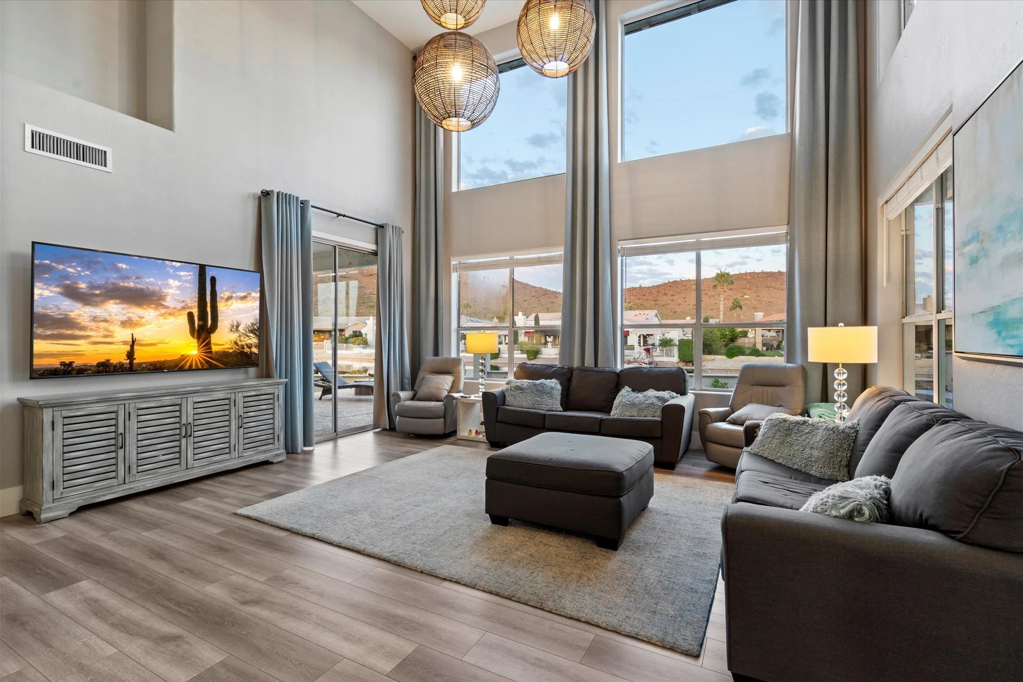 Bright living room with large windows showing a mountain view, featuring a gray sofa, an armchair, a TV on a console, and hanging wicker light fixtures.