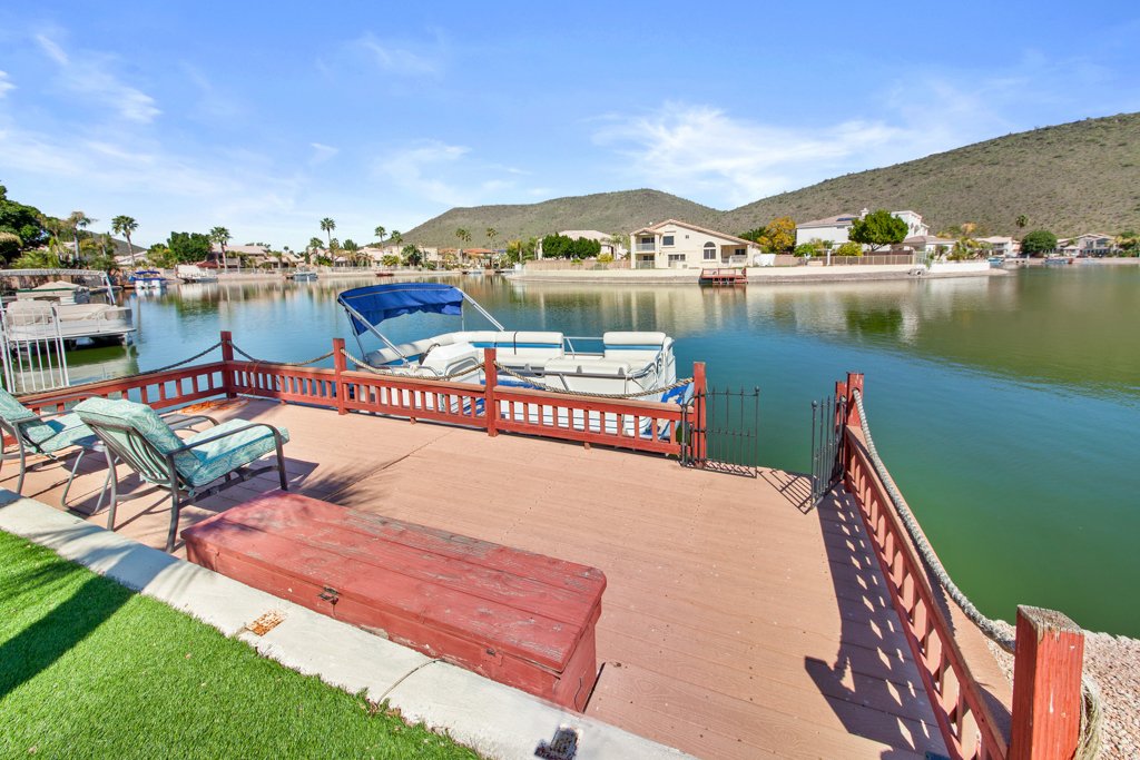 A waterfront patio with a wooden deck, lounge chairs, and a boat with a canopy moored in a calm water canal, with houses and hills in the background under a clear blue sky.