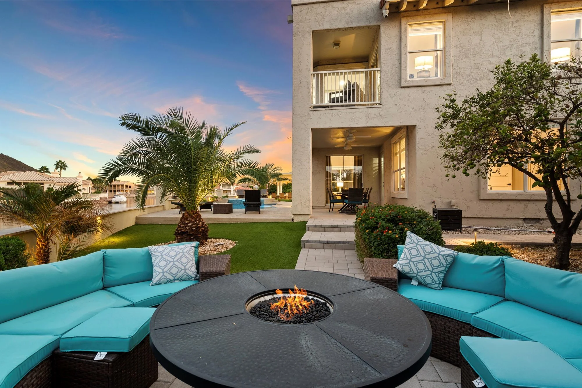 Outdoor patio at twilight with a round fire pit in the center, a curved turquoise sectional sofa with decorative pillows, palm trees, a swimming pool, and a multi-story beige house with lit windows.