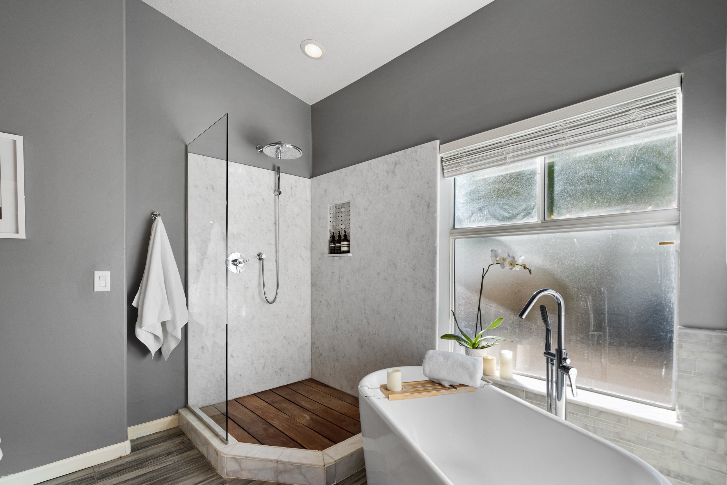 Modern bathroom with a gray wall, a walk-in shower with glass door and a rainfall showerhead, wooden floor mat, bathtub with candles and a plant near the window.