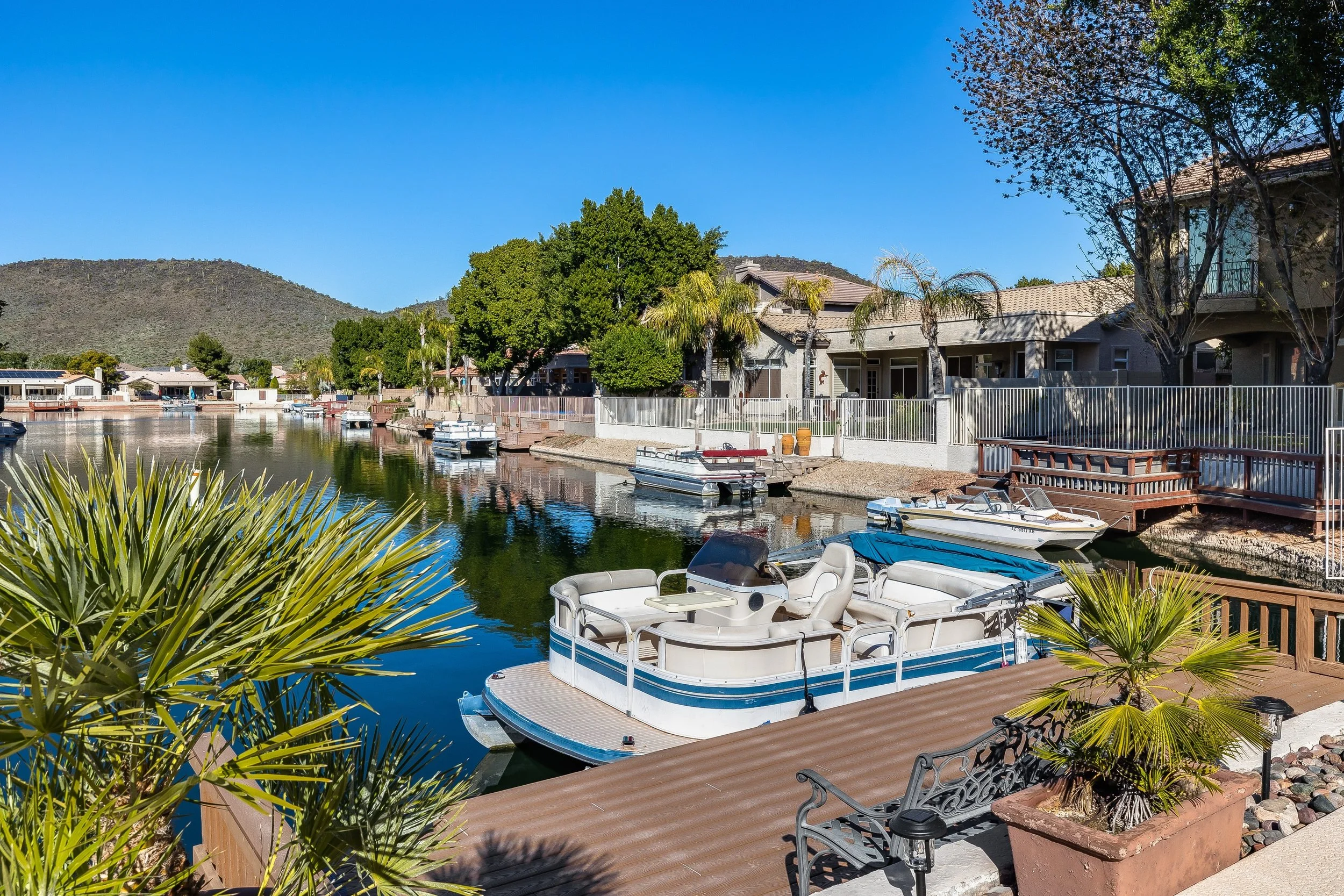 A canal with boats docked along the side, residential houses with patios, and lush green trees under a clear blue sky in a sunny setting.