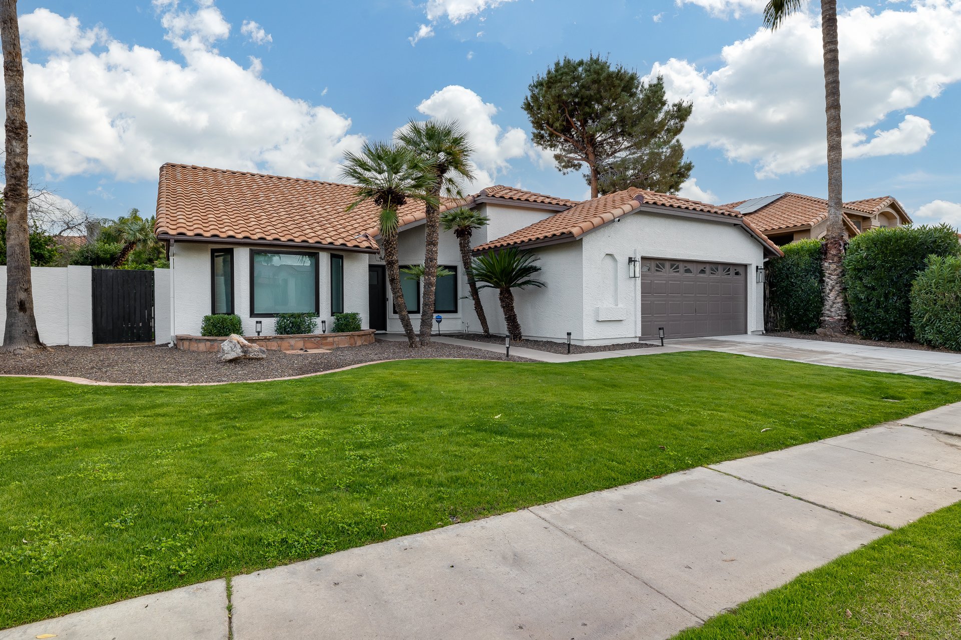 Front yard of a modern white house with a tiled roof, two palm trees, a lush green lawn, sidewalk, driveway, and a black gate on the left side.