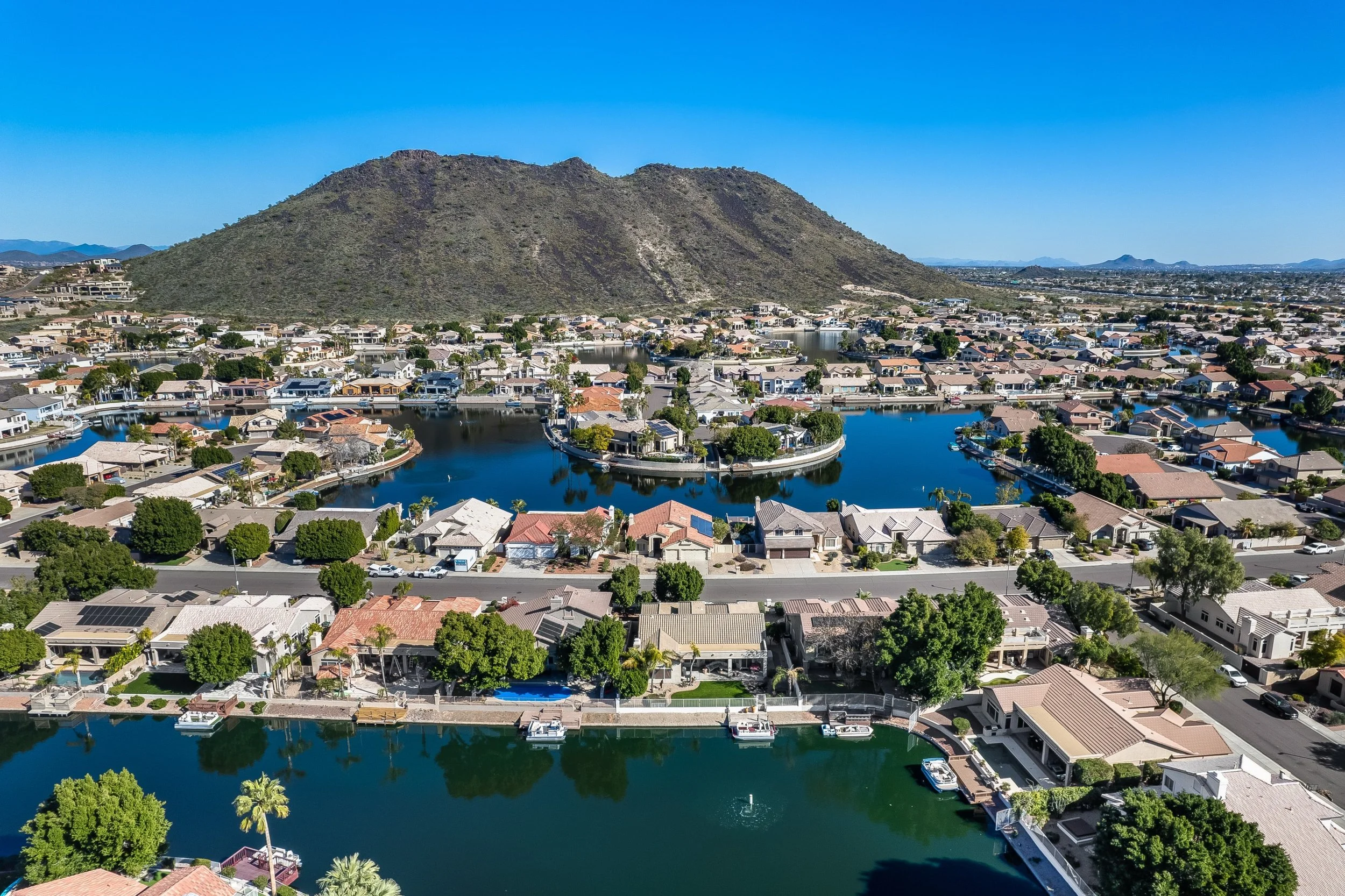 Aerial view of a suburban neighborhood with houses, water canals, and trees, with a mountain in the background under a clear blue sky.