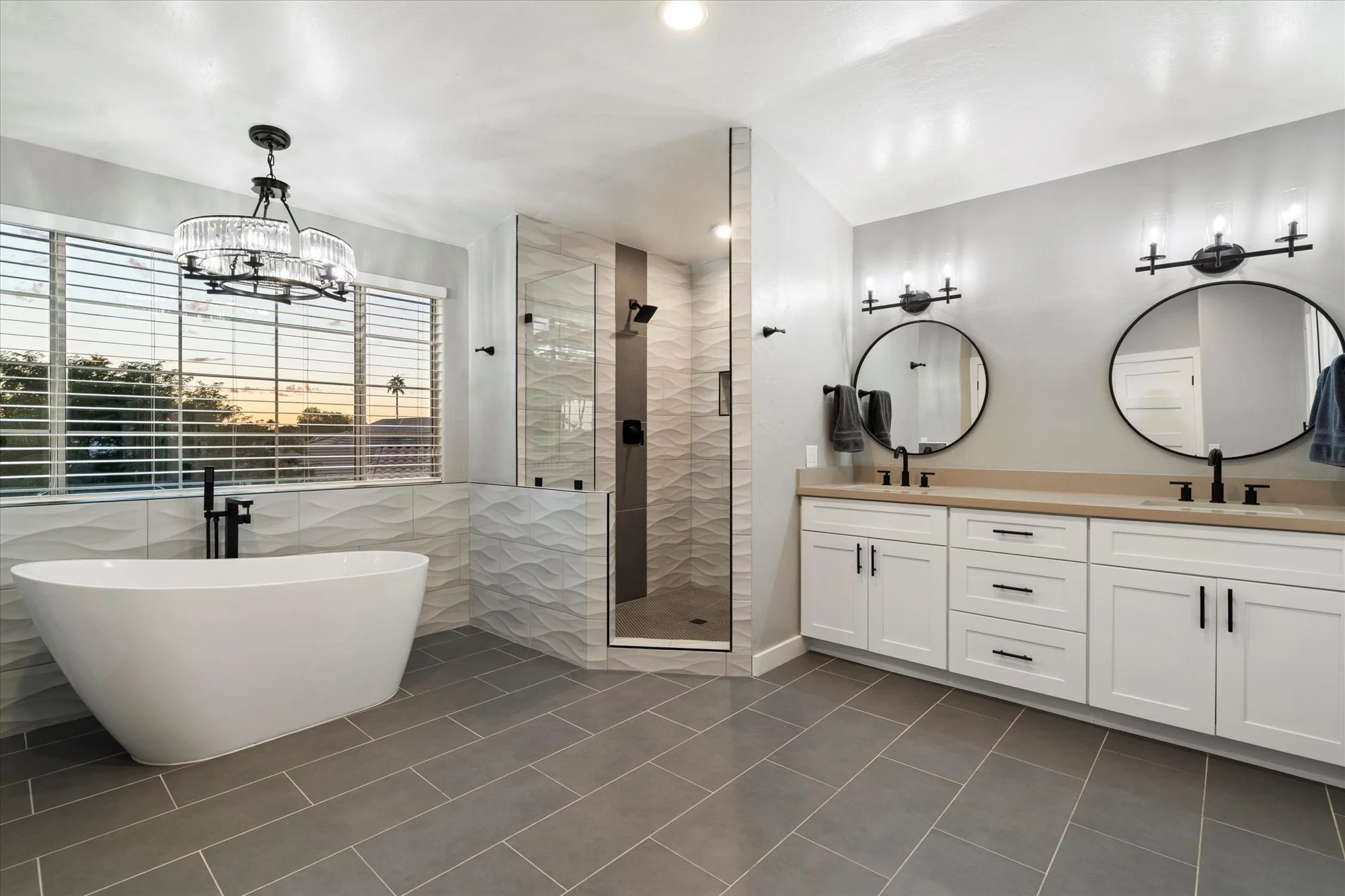 Modern bathroom with freestanding bathtub, walk-in shower with textured tiles, double vanity with large mirrors, black fixtures, and a large window showing sunset.