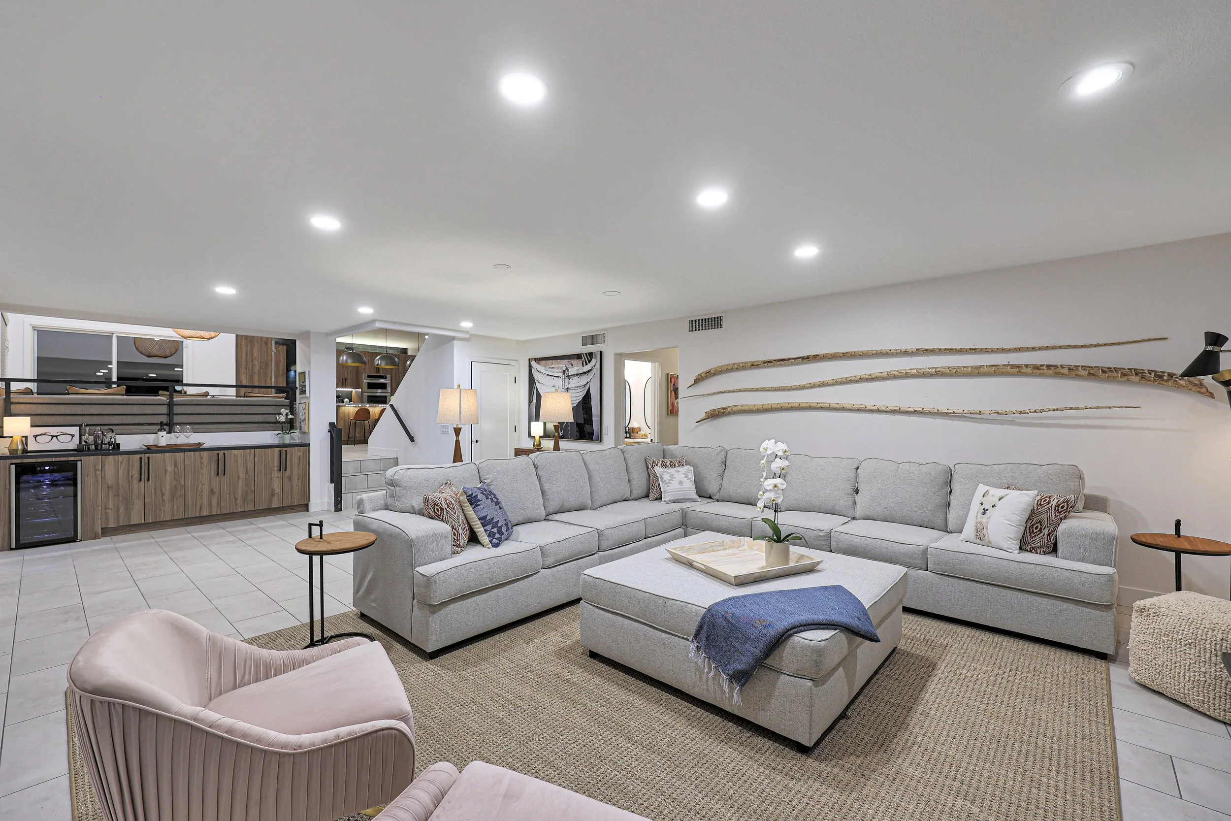 A spacious living room with a large L-shaped light gray sofa decorated with patterned pillows and a dark blue blanket. A smaller pink upholstered armchair is in the foreground. The room features a tiled floor, a neutral color palette, and decorative 