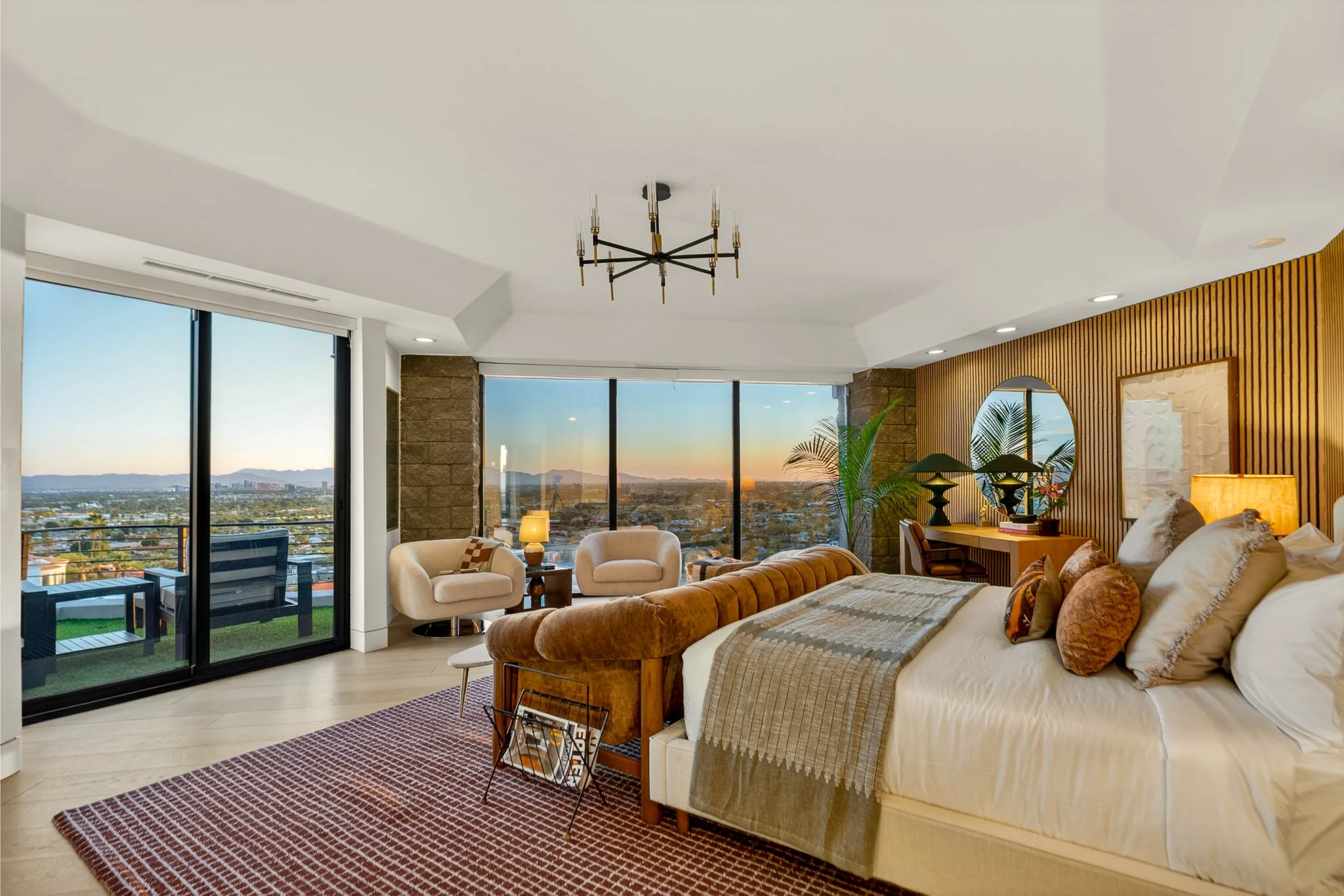 Living room with large windows showing a city and mountain view, beige and brown furniture, modern chandelier, and decorative items on a wooden accent wall.