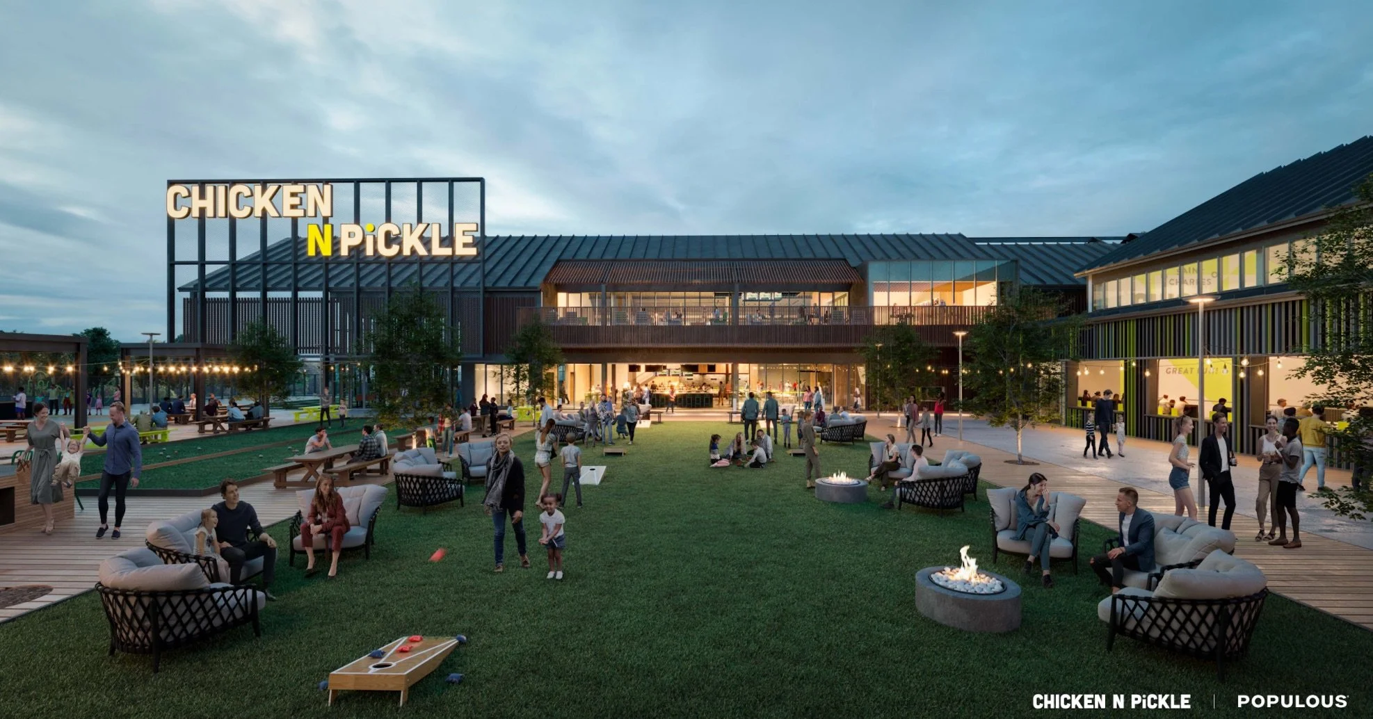 A lively outdoor area in the evening with many people gathered, sitting, standing, and walking around. There are illuminated buildings, a fire pit, benches, and a grassy space with children playing. The prominent sign on the building reads, 'CHICKEN N PICKLE.'