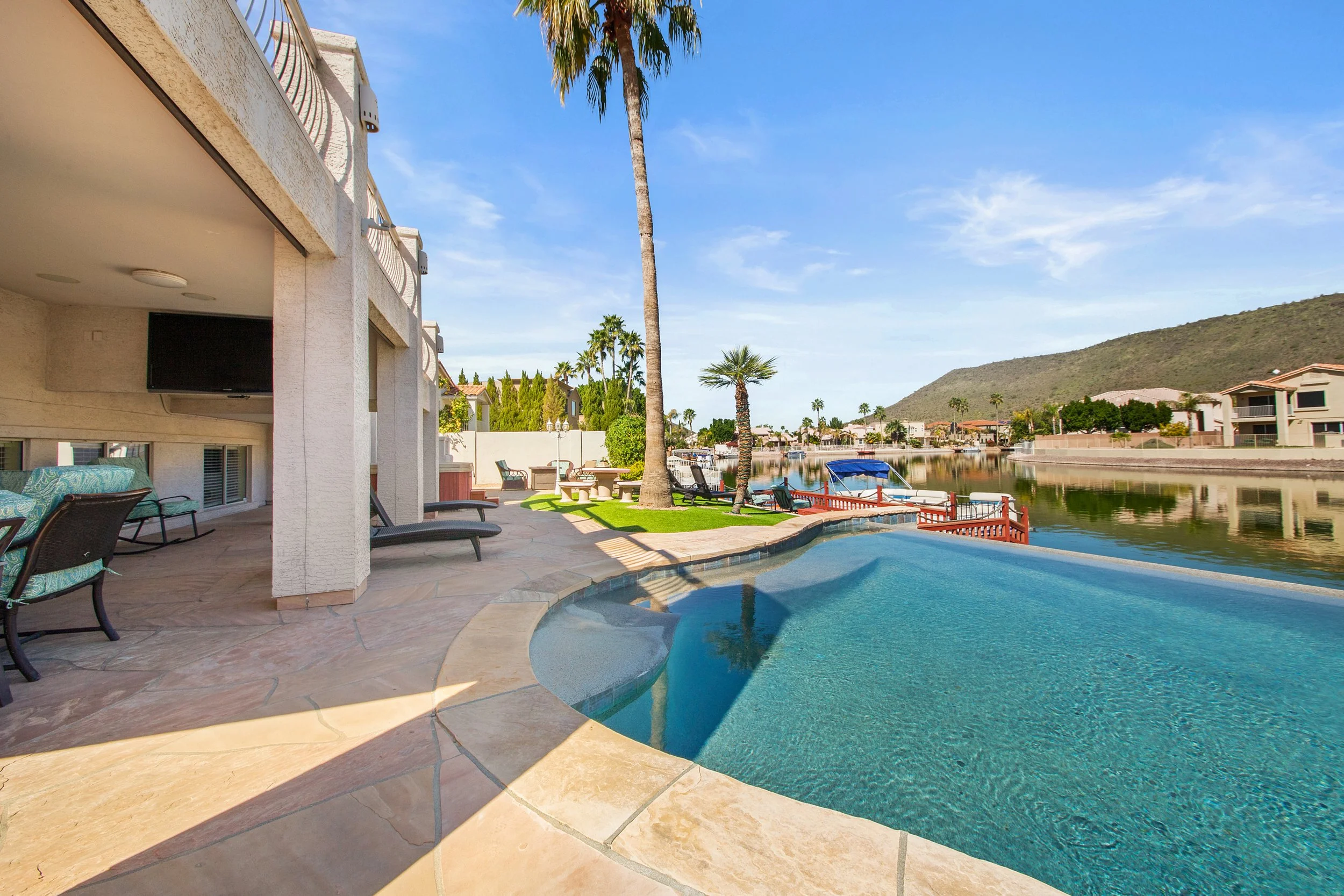 A backyard with a swimming pool and a view of a canal with boats, surrounded by palm trees and houses with mountains in the background on a sunny day.