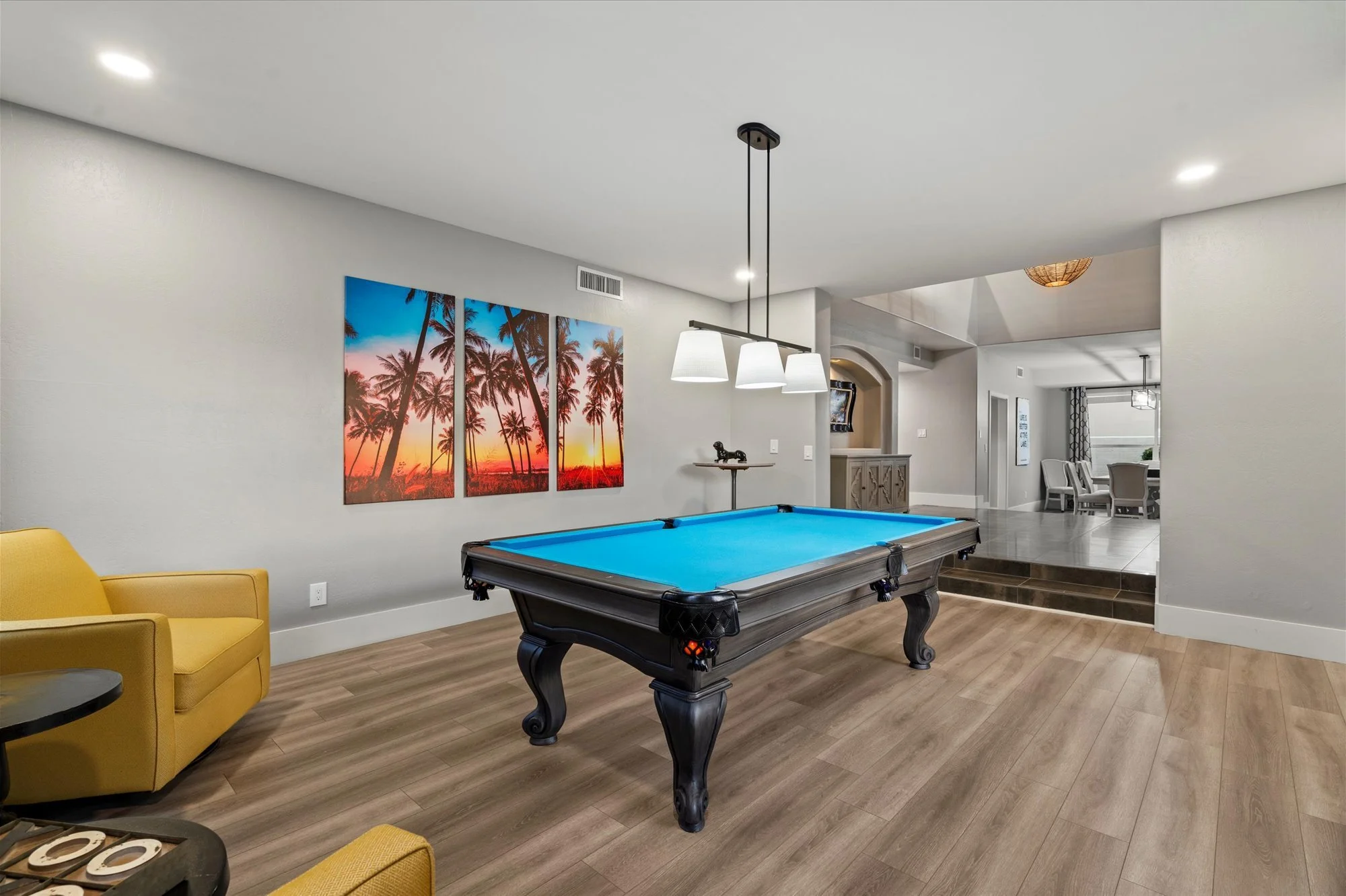 Living room with a pool table, yellow armchair, wall art of palm trees during sunset, and dining area in the background.