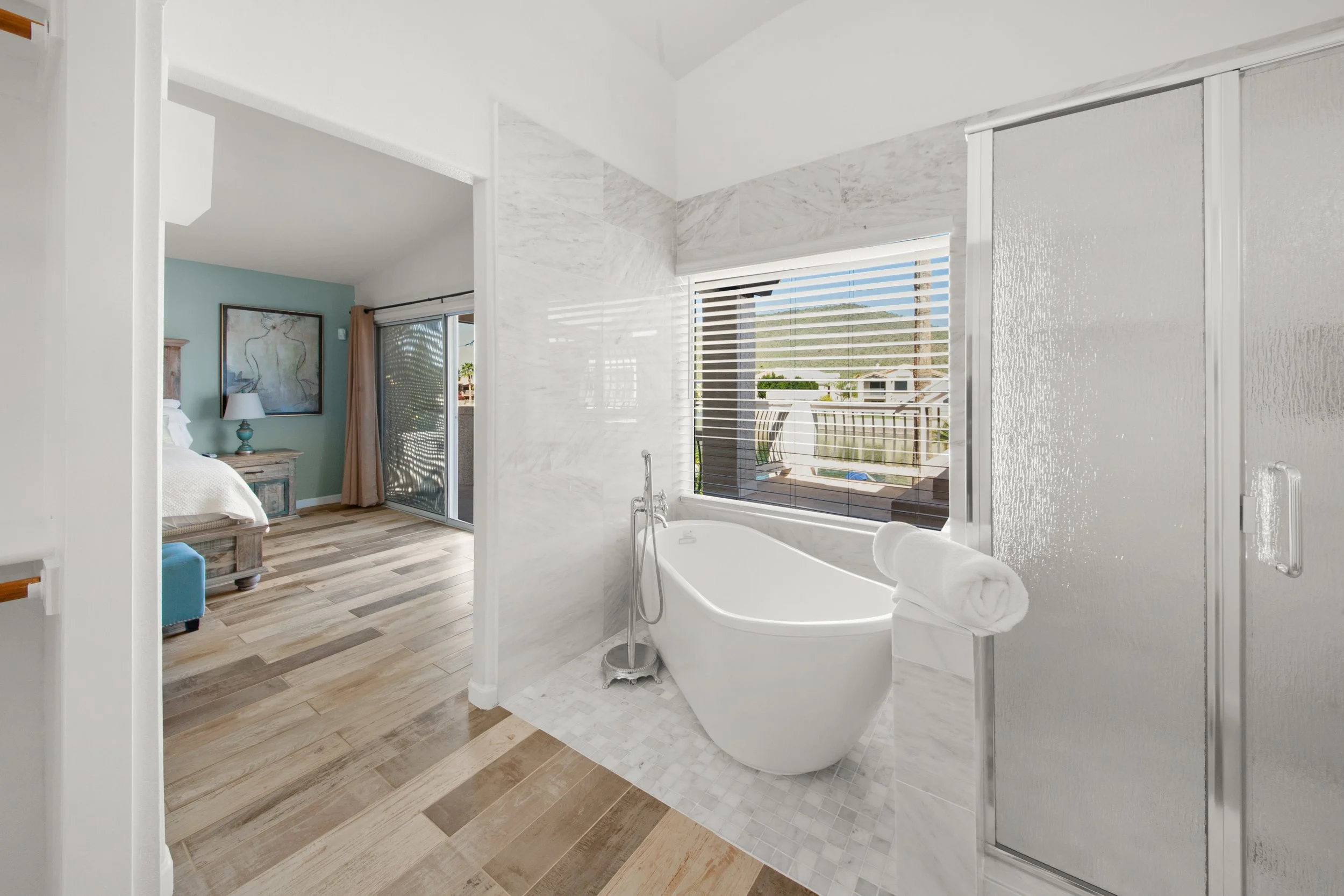 Modern bathroom with white bathtub, large window with city view, and bedroom with artwork and natural light.
