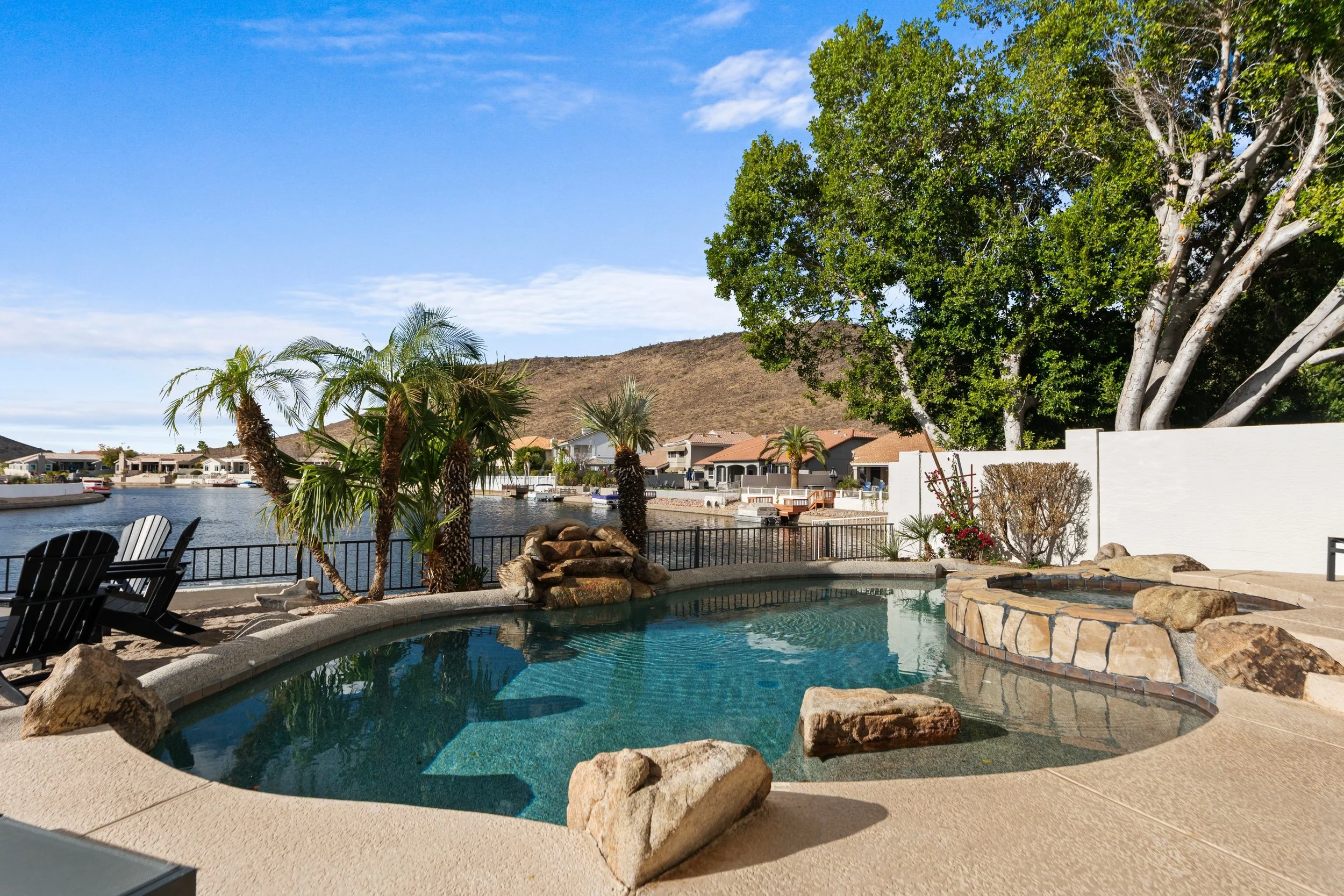 A backyard with a swimming pool surrounded by rocks, palm trees, and a black metal fence, overlooking houses and a hill in the background.