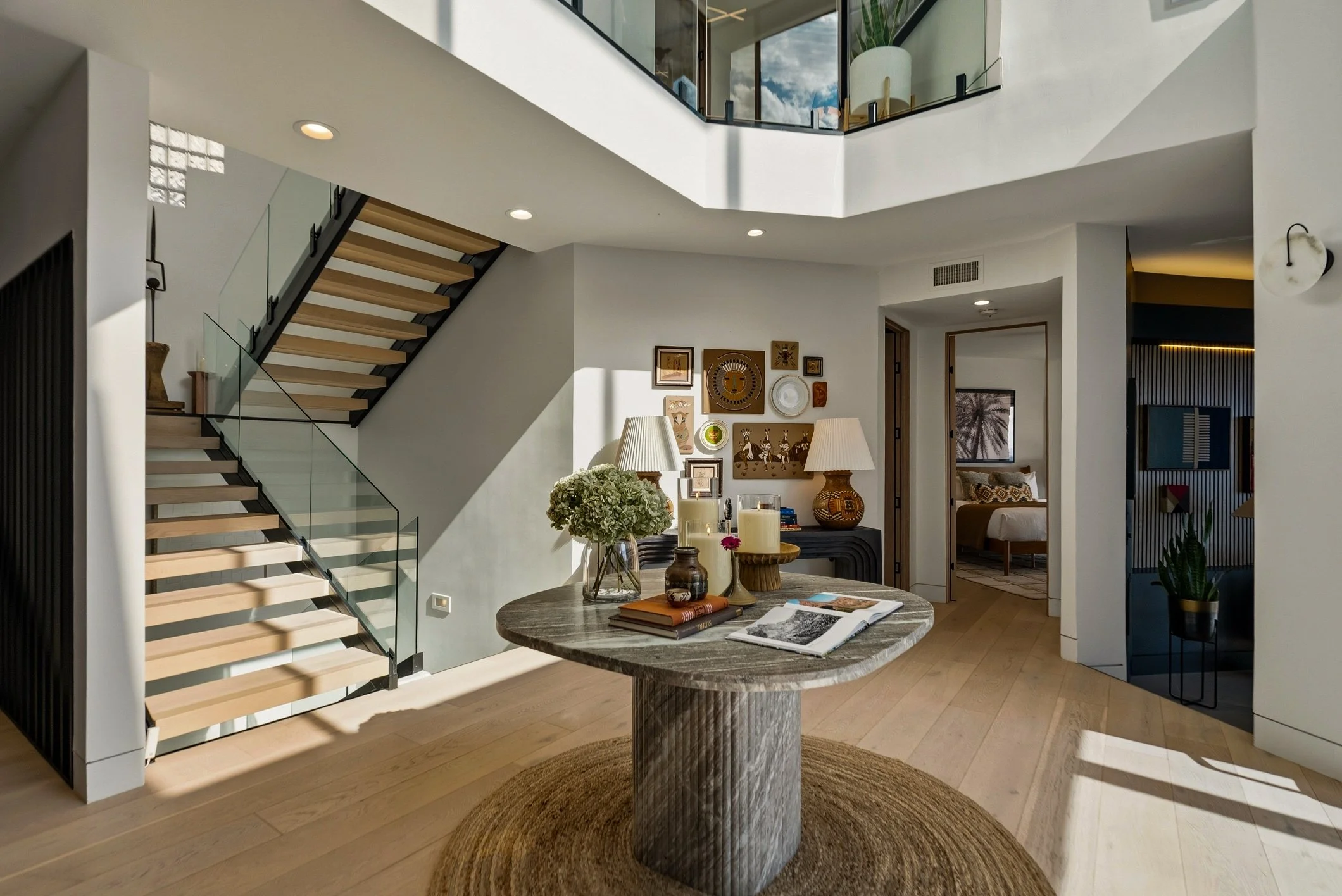 Interior view of a modern, open-concept living space with a staircase, wall art, and a dining table with decorative items. Natural light from the windows illuminates the room.