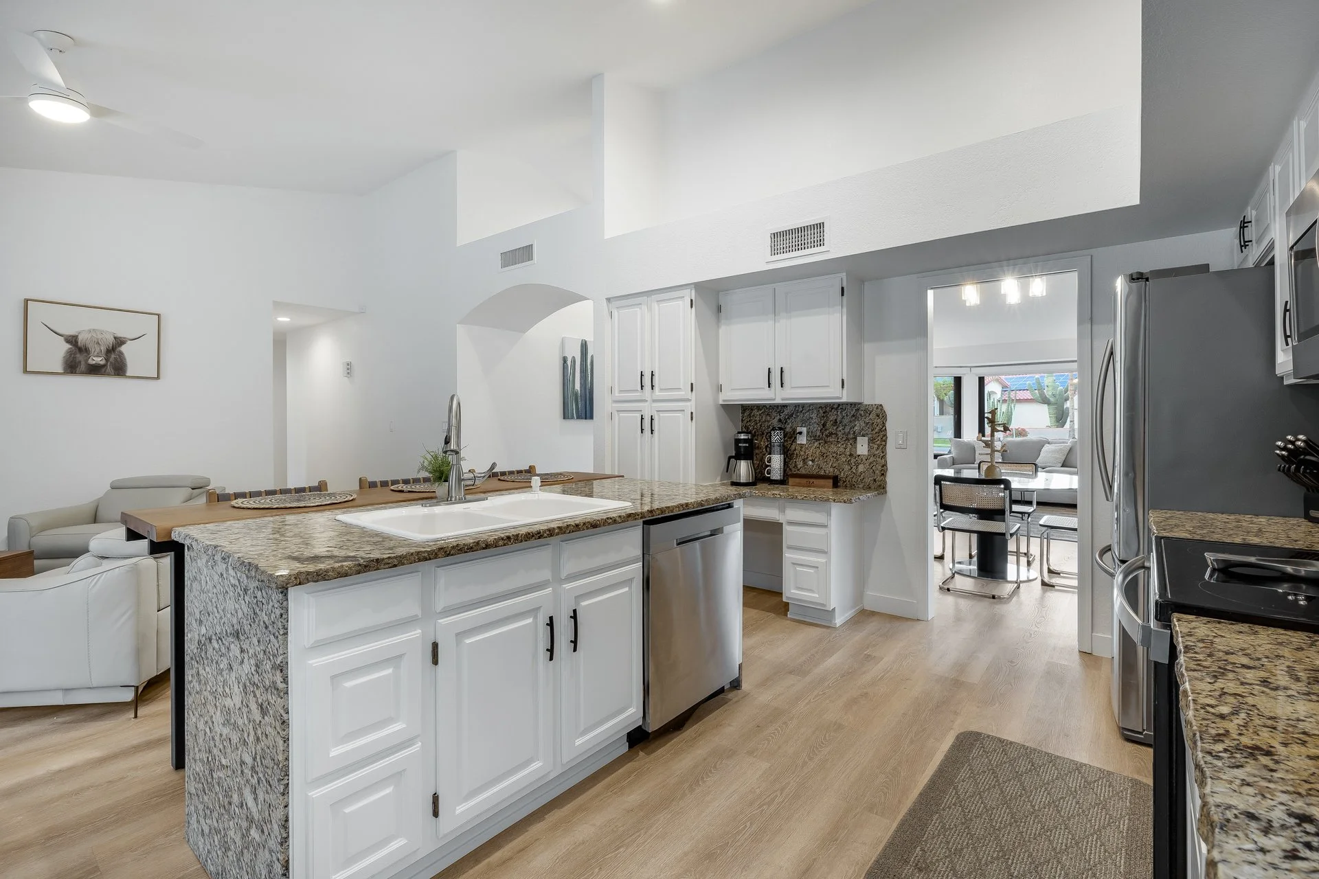 Open-concept white kitchen with granite countertops, stainless steel appliances, and a view into a dining area with seating and a large window.