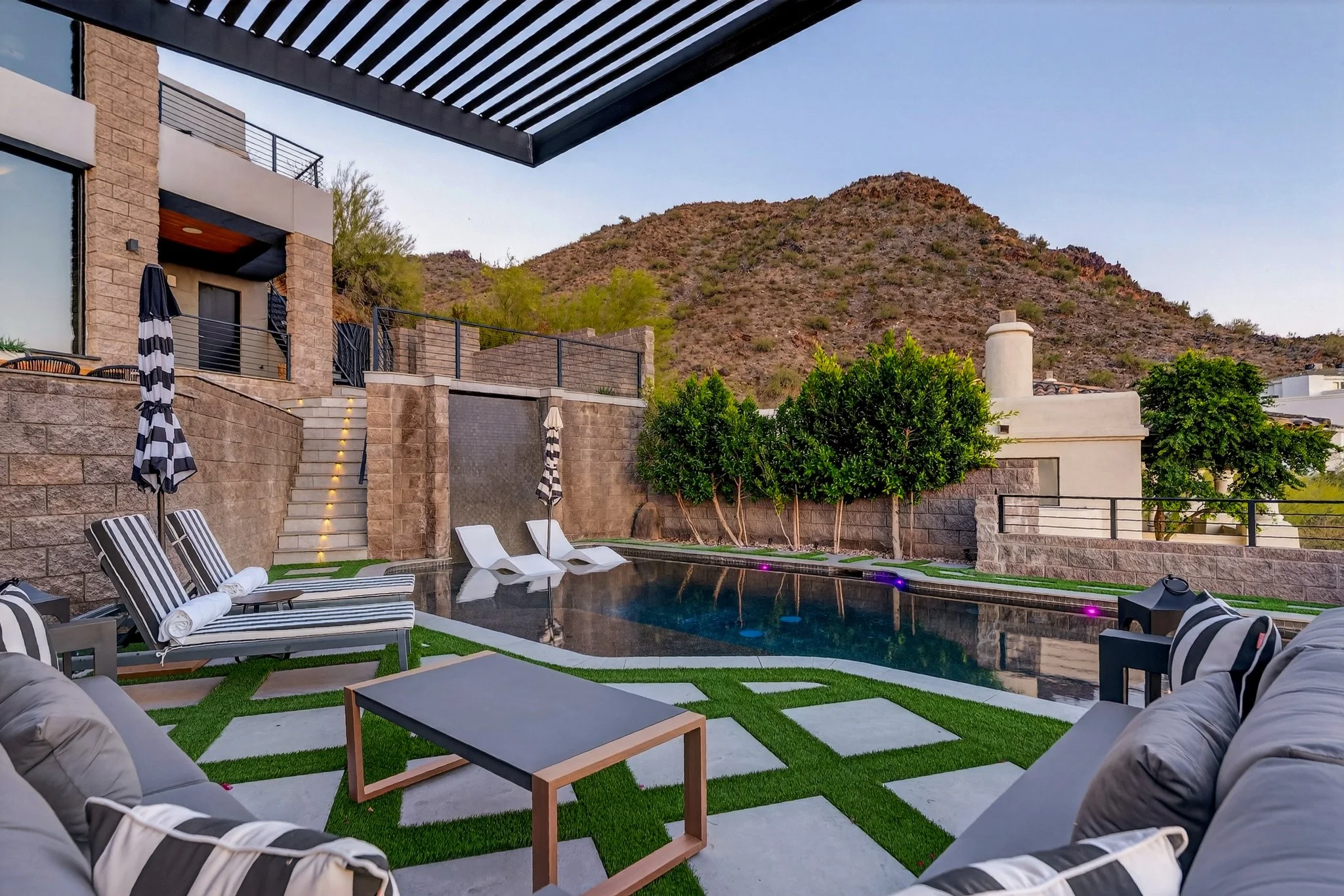 View of a modern outdoor backyard with a swimming pool, lounge chairs with striped cushions, and patio furniture surrounded by trees and a mountain in the background.