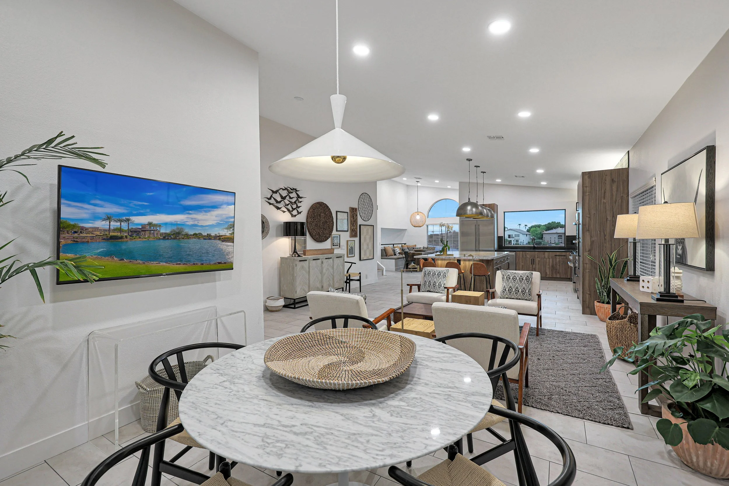 Open concept living room and kitchen with white walls, pendant lights, artwork on the wall, a flat-screen TV showing a lake scene, dining area with a marble table and black chairs, and plants surrounding the space.