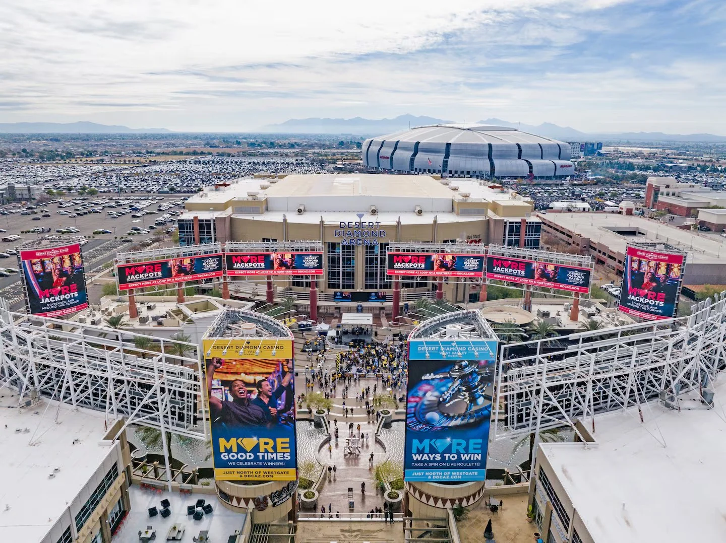 Aerial view of Desert Diamond Arena and surrounding parking lots, with digital billboards displaying advertisements and a crowd of people entering the arena.