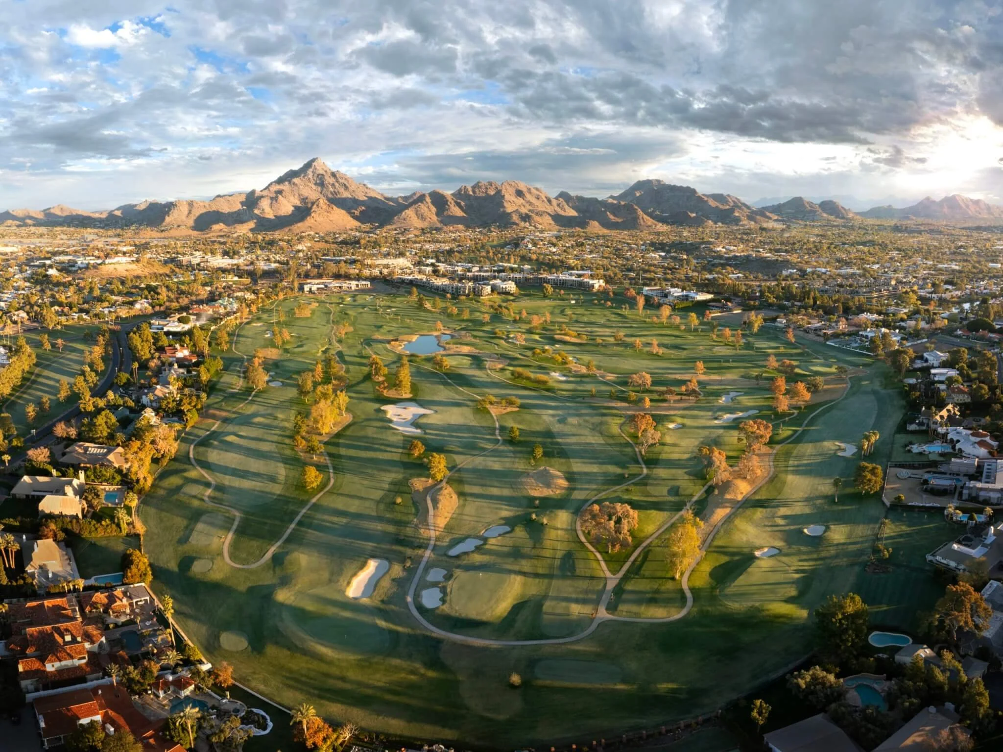 Aerial view of a city with mountains in the background, green golf course in the foreground, scattered houses, and partly cloudy sky.