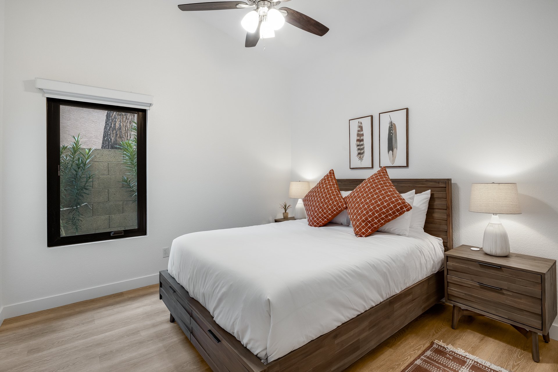 A bedroom with white walls, a large bed with white linens, two orange patterned pillows, and wooden furniture including two nightstands with lamps. There are two framed feather artworks above the bed, and a window with white blinds showing some green
