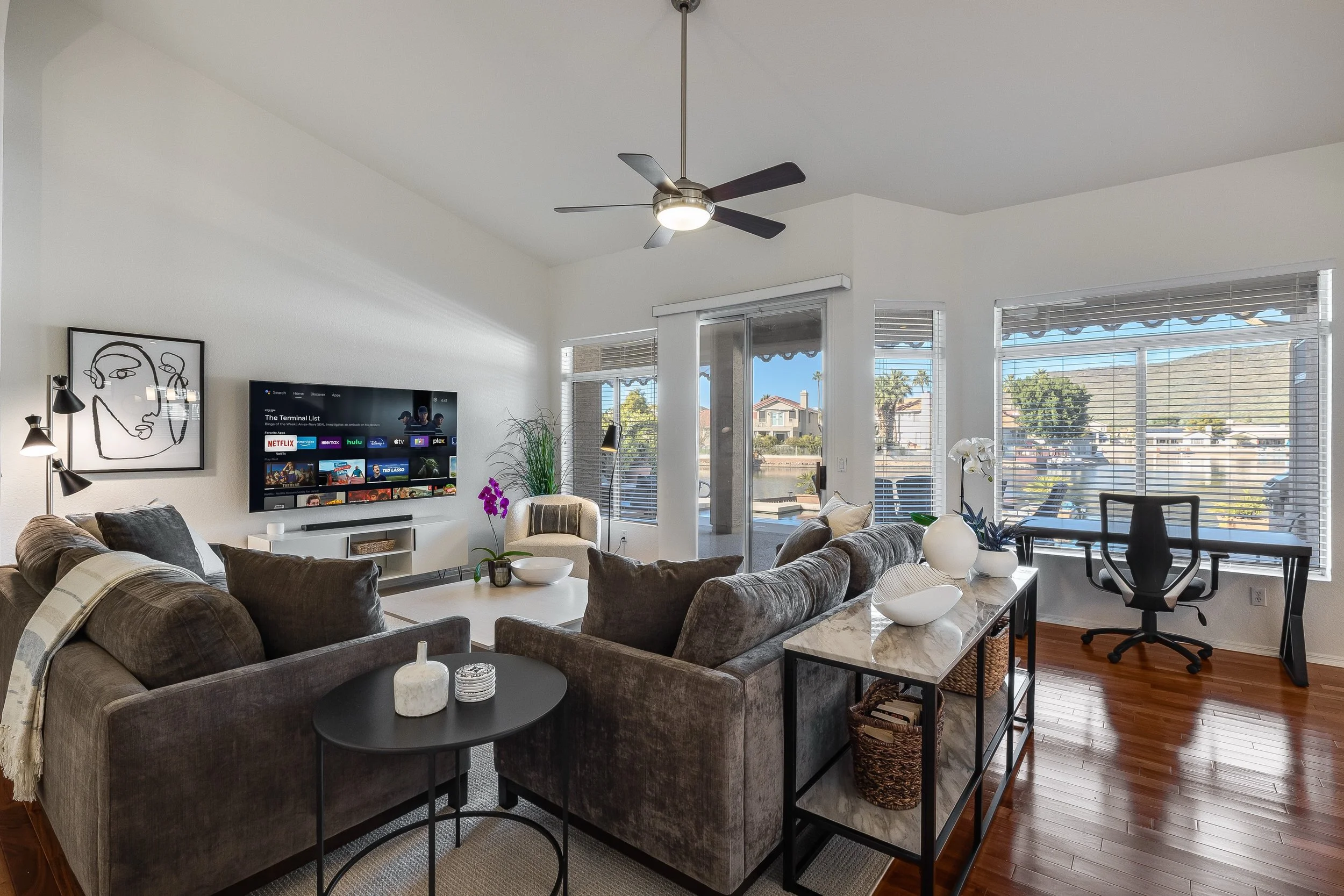 A bright living room with large windows, a ceiling fan, a mounted TV, and a variety of furniture including a gray sectional sofa, a white coffee table, and a black side table with decorative items. There are plants and artwork on the walls, and the r