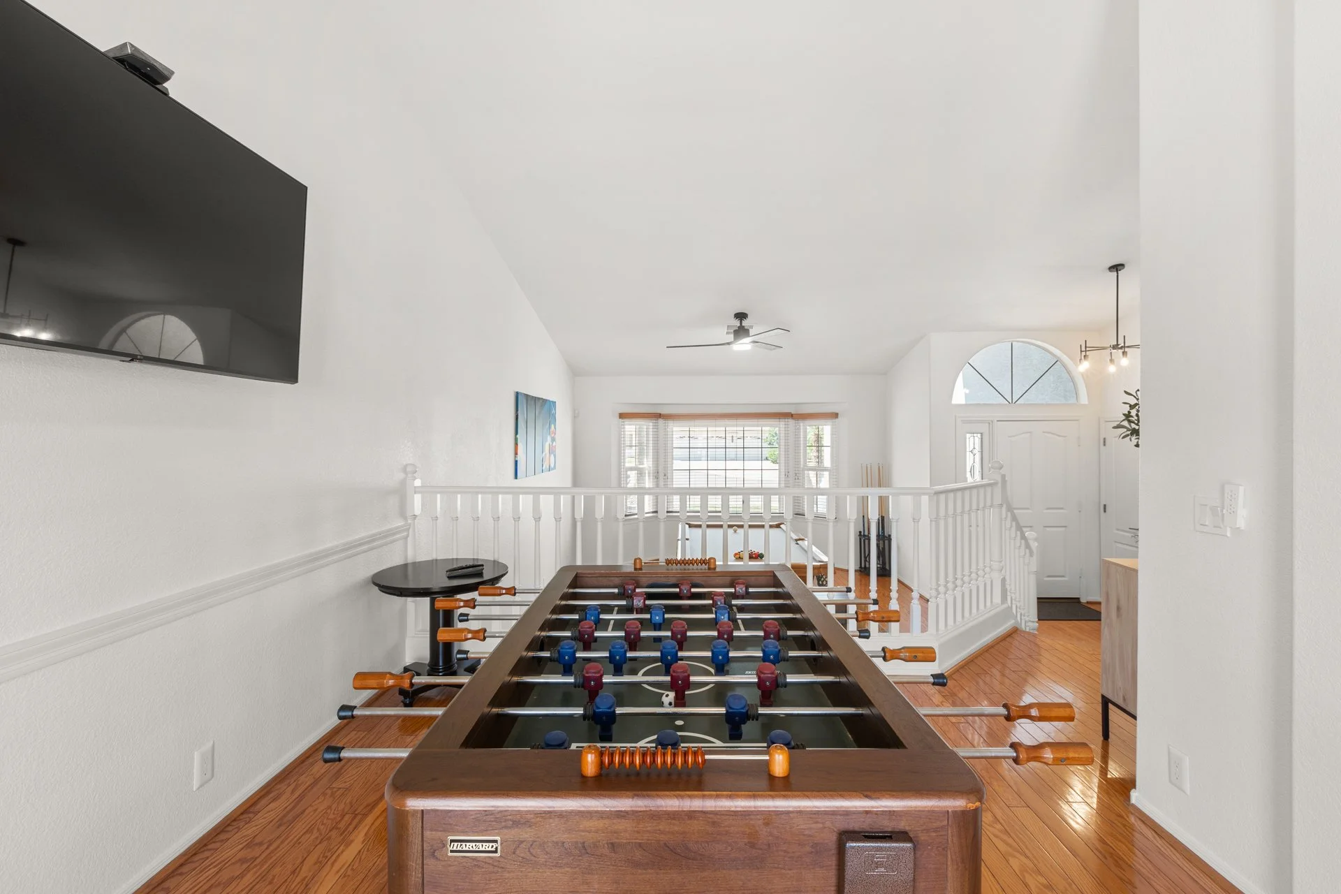 Interior view of a bright living room with a foosball table, a wall-mounted TV, a ceiling fan, and windows with blinds, featuring white walls and hardwood floors.