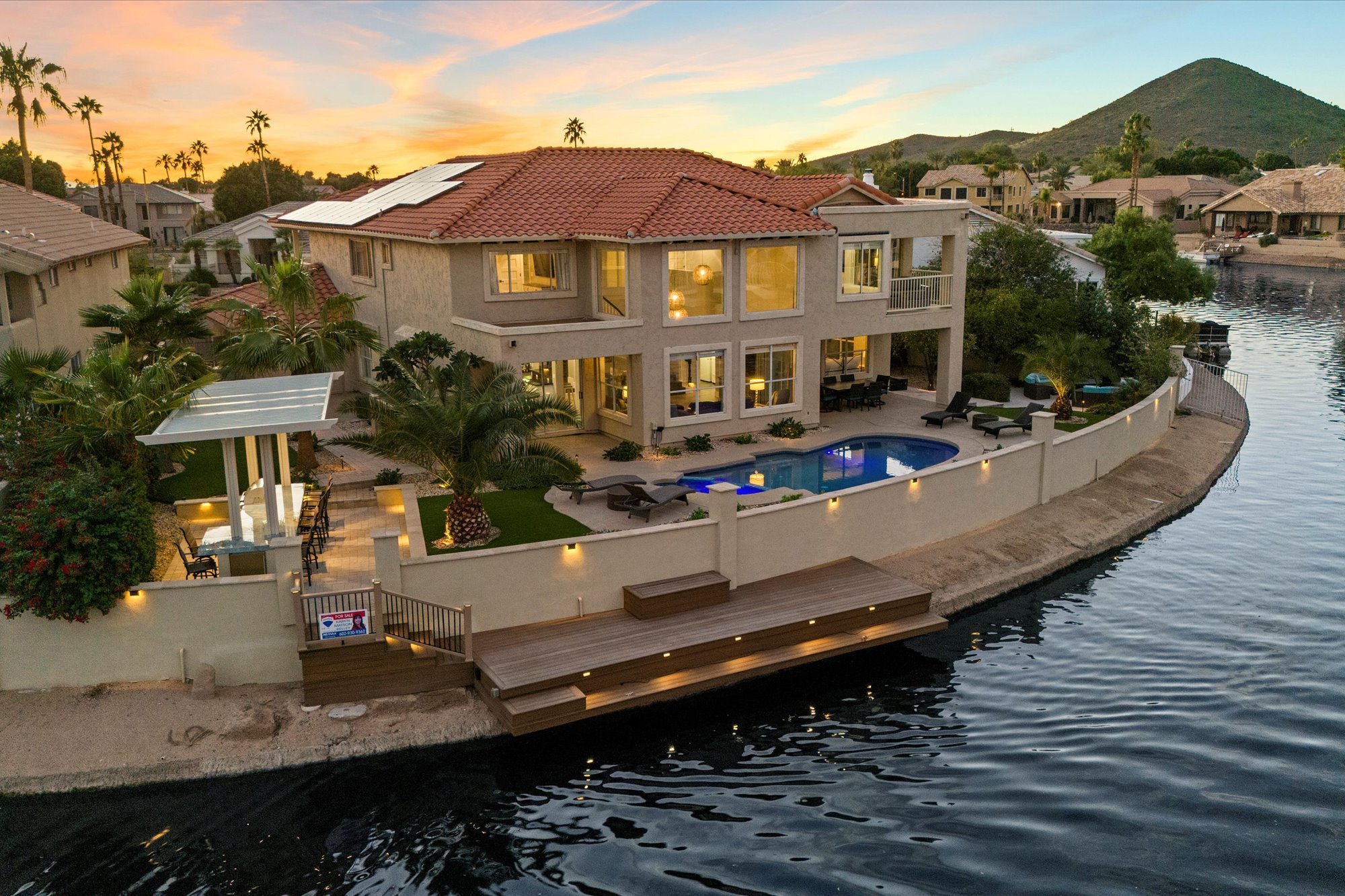 Luxury waterfront house with a red tile roof, large windows, a swimming pool, and a dock in a neighborhood with palm trees at sunset.