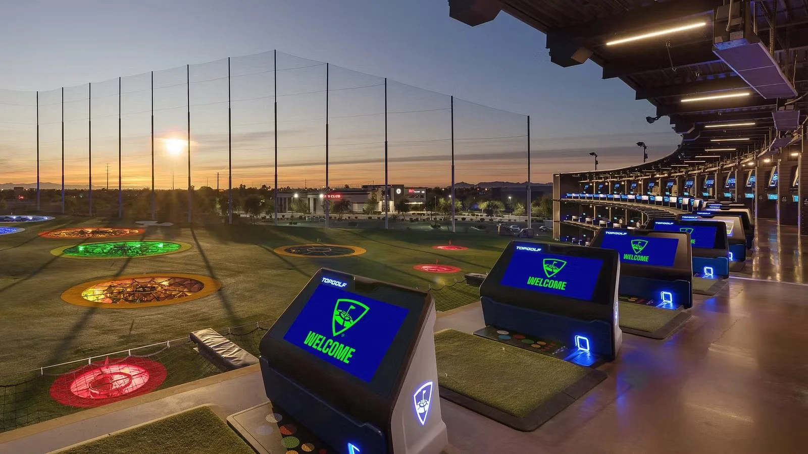 An outdoor mini-golf driving range at sunset with multiple golf simulators and illuminated holes, overlooking a fenced golf course.