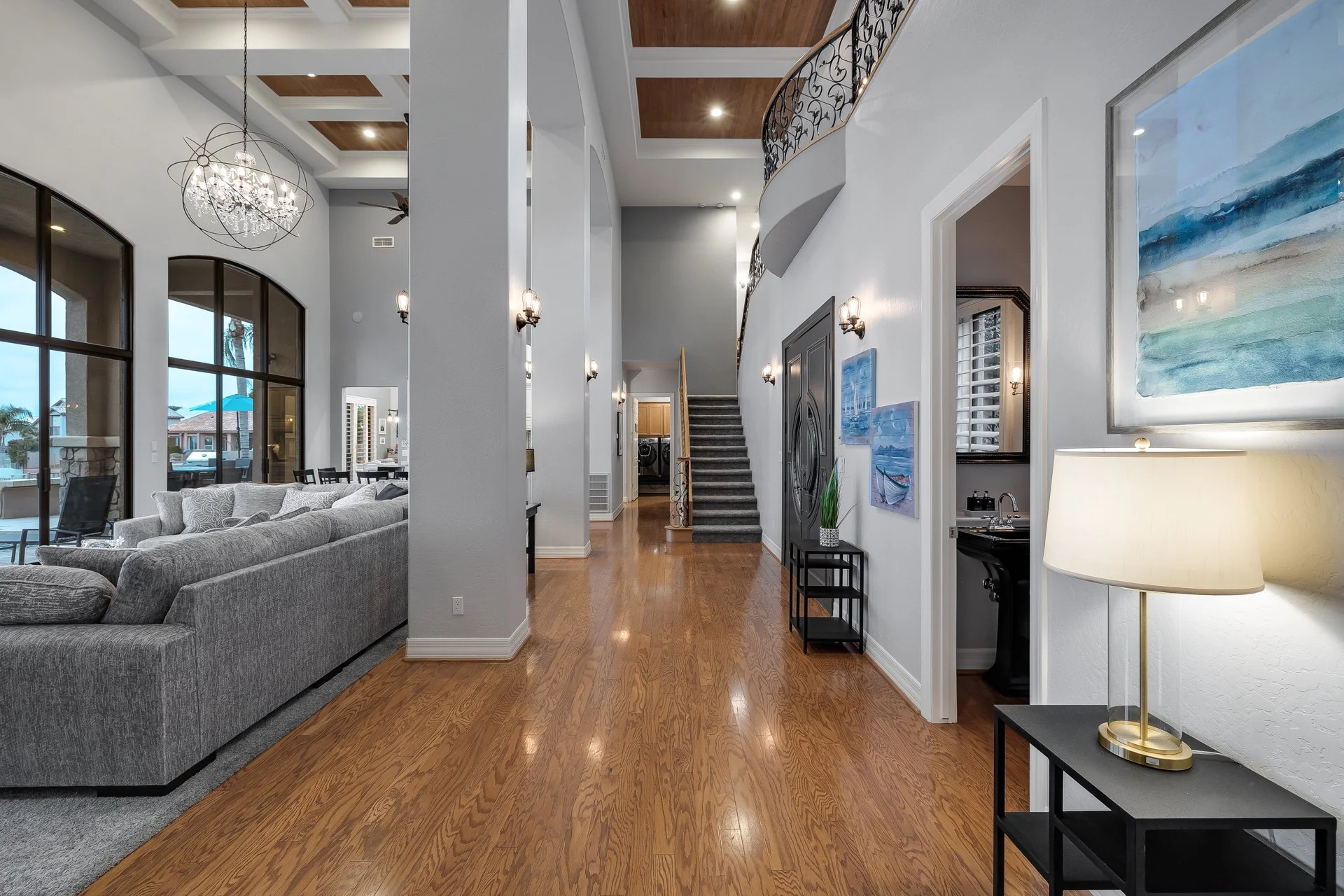 Interior view of a spacious, modern home with high ceilings, hardwood floors, a curved staircase, and large windows, including a living area with a gray sofa and artwork on the walls.