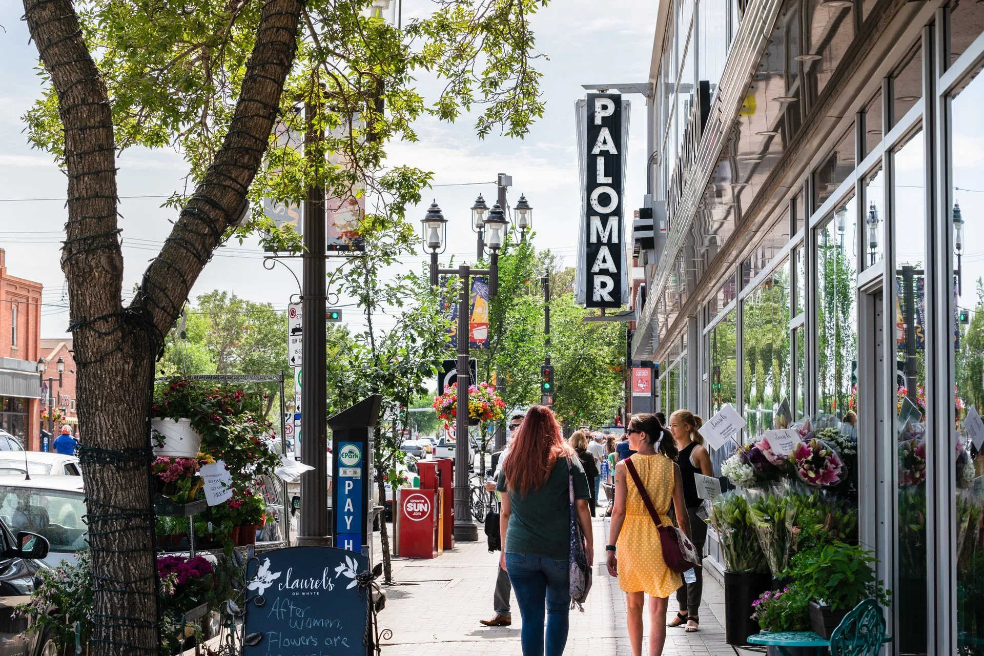 Street scene with pedestrians, storefronts, and outdoor plants. A sign reads 'PALOMAR'. Trees line the sidewalk, and a pay parking meter is visible.
