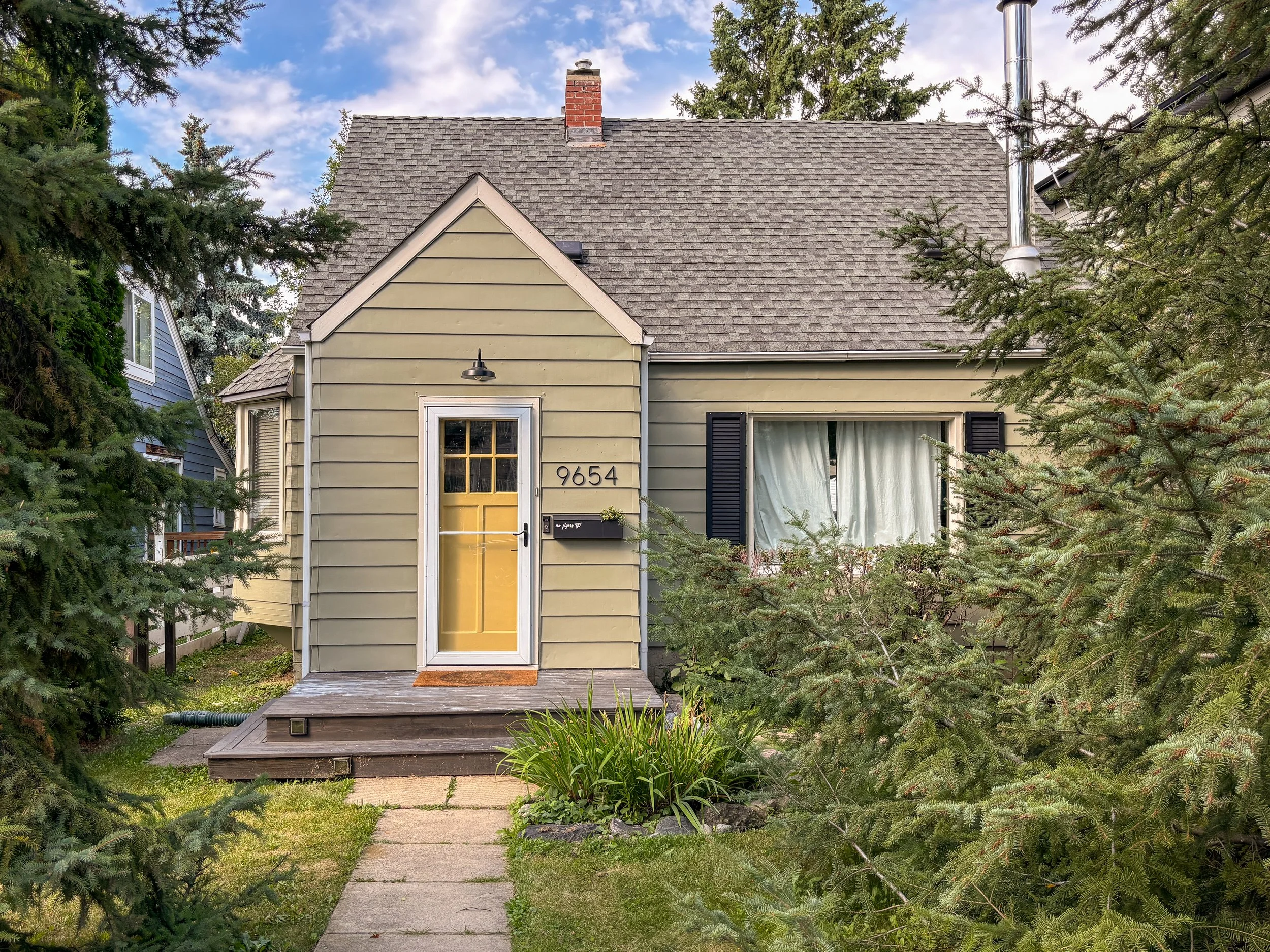 Front view of a small house with a green exterior, yellow door, a small porch, and a window with black shutters surrounded by greenery and trees.