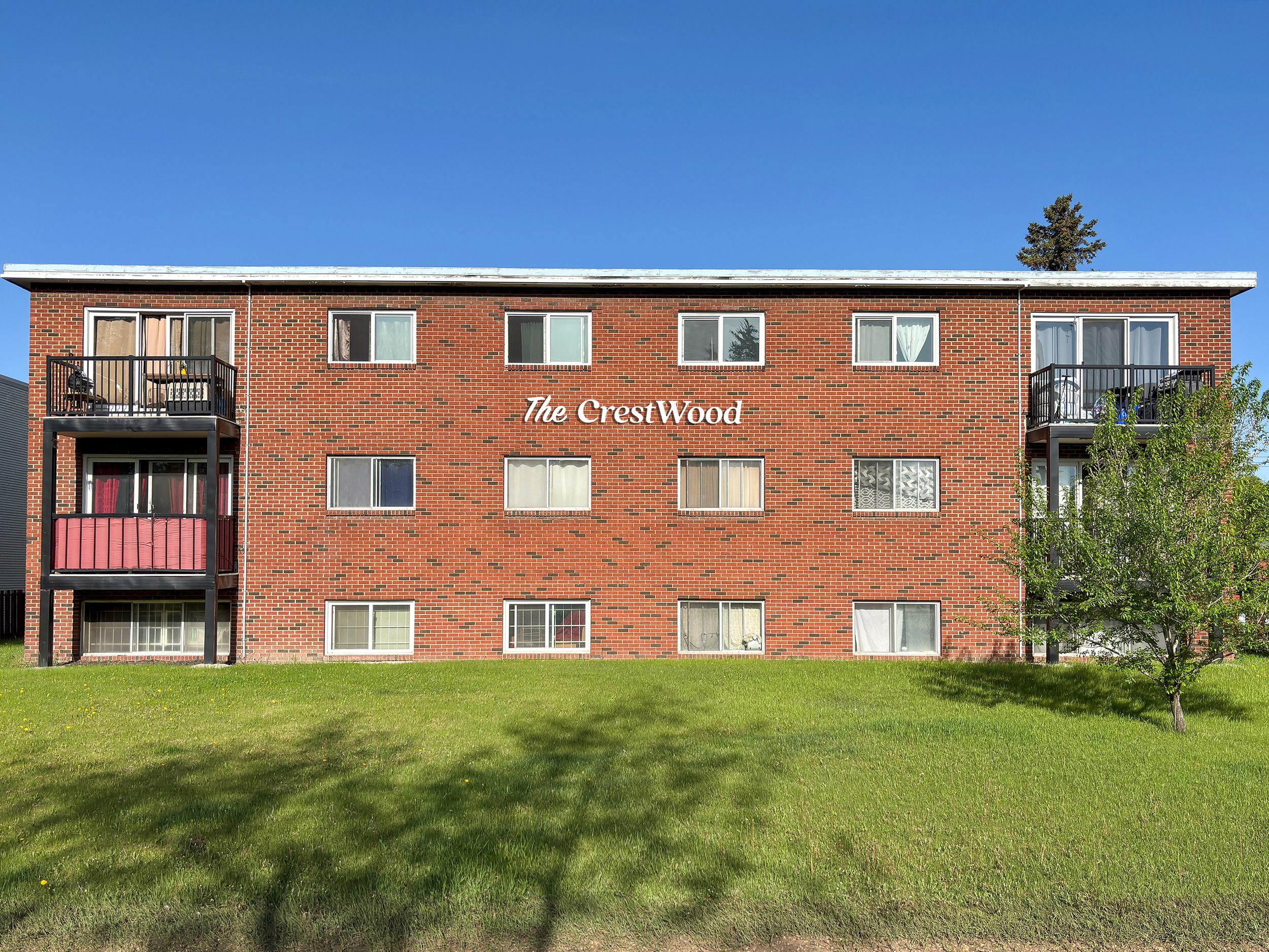Red brick apartment building called 'The CrestWood' with black balconies and a green lawn in front under a clear blue sky.