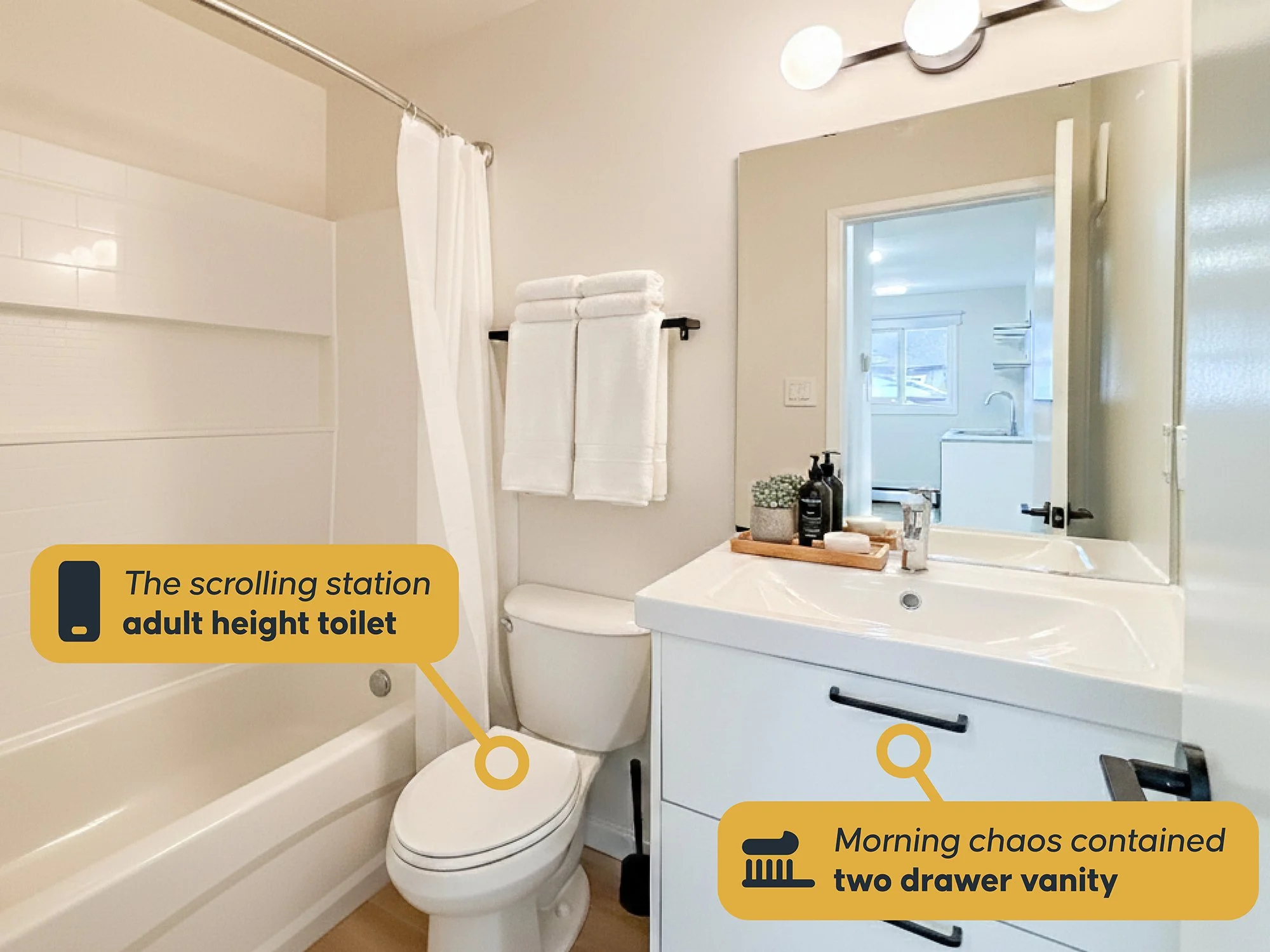 A bathroom with a bathtub, a toilet, and a vanity with a large mirror. Towels are hanging on a rack behind the toilet, and various toiletries are on the vanity tray. There is natural light coming through a nearby window.