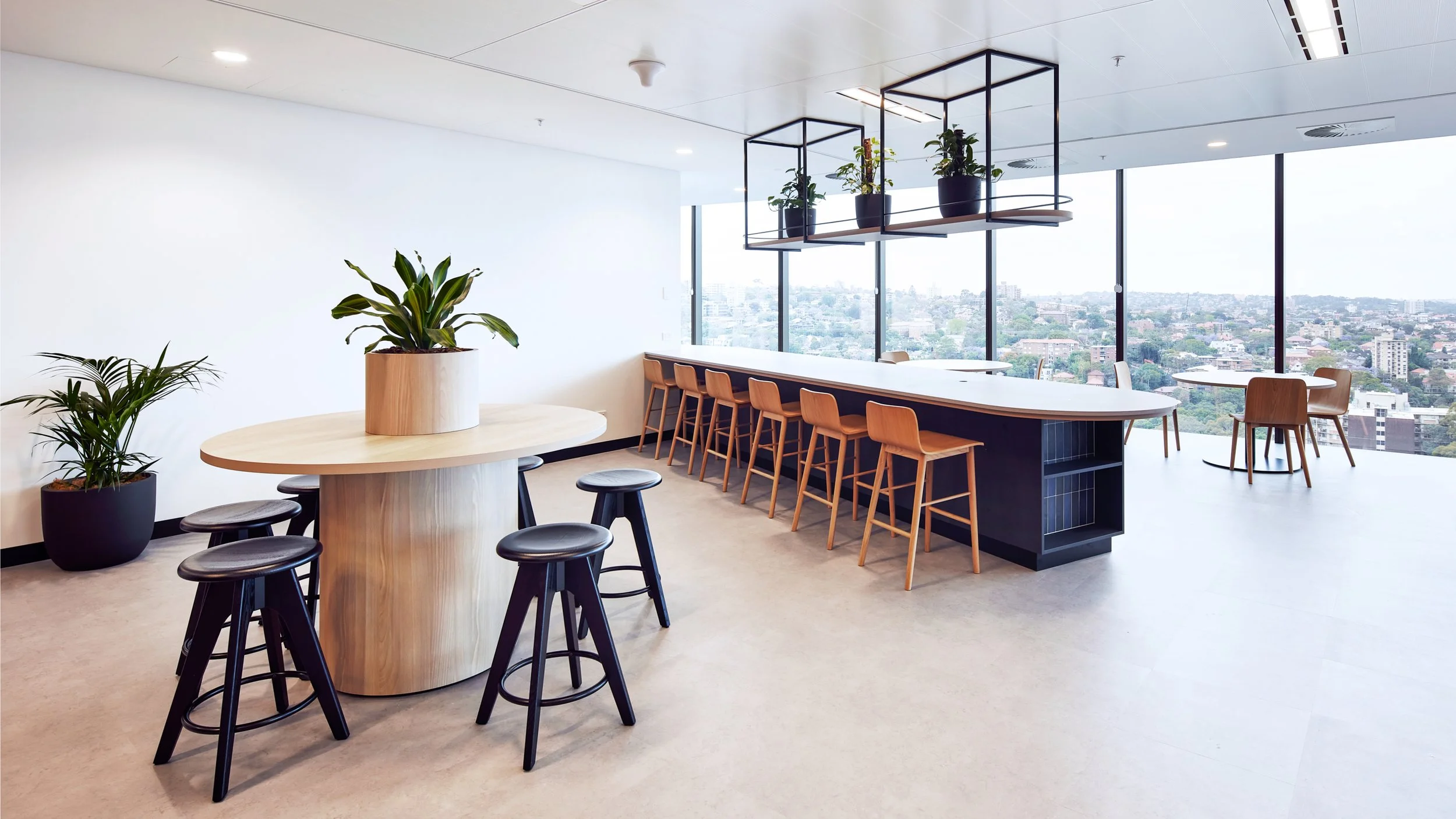 An stylish kitchen area features a two timber tables and some plants that are fixed closed to the ceiling, in the background glass panels overlook the landscape