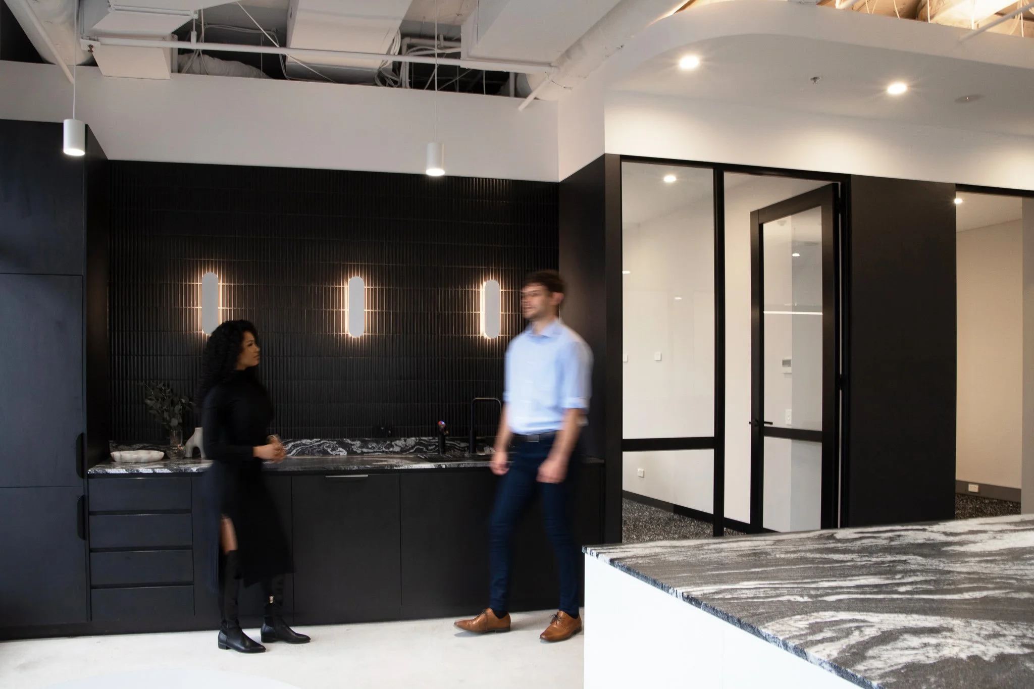 An elegant kitchen piece in a dark pallete is seen behind two workers talking