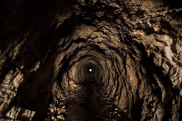 An image looking down of a dark, rocky tunnel towards a small light at the end. The rocky walls are brown and roughly textured.